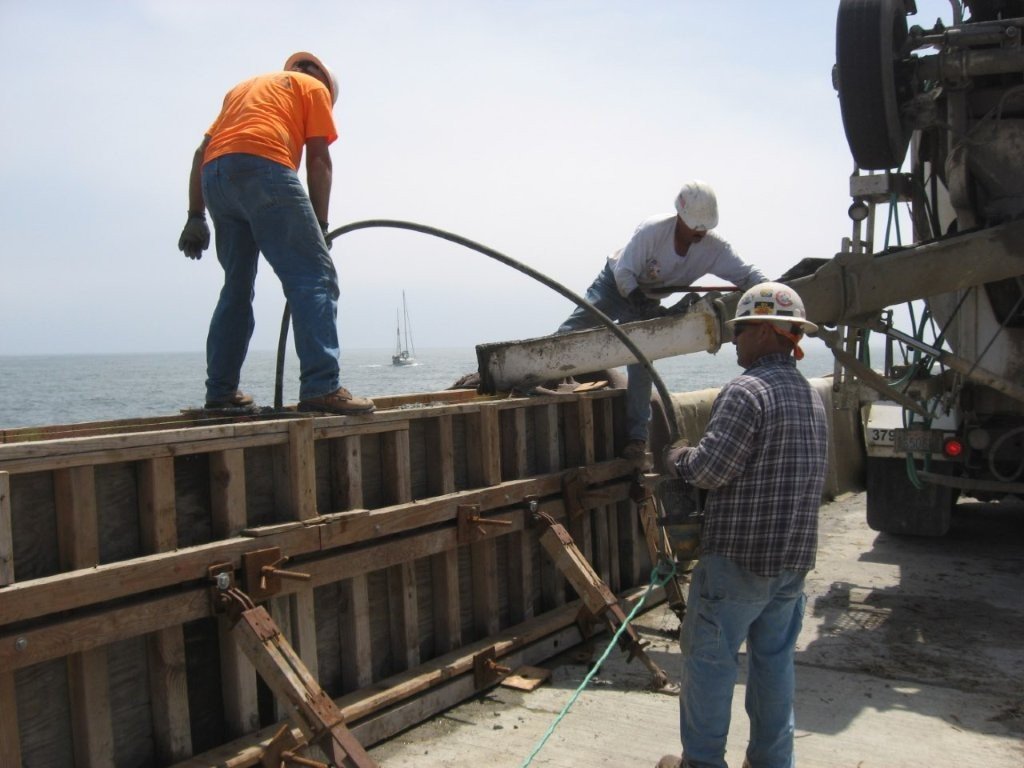 Workers are pouring concrete into a formwork on a construction site by the ocean.