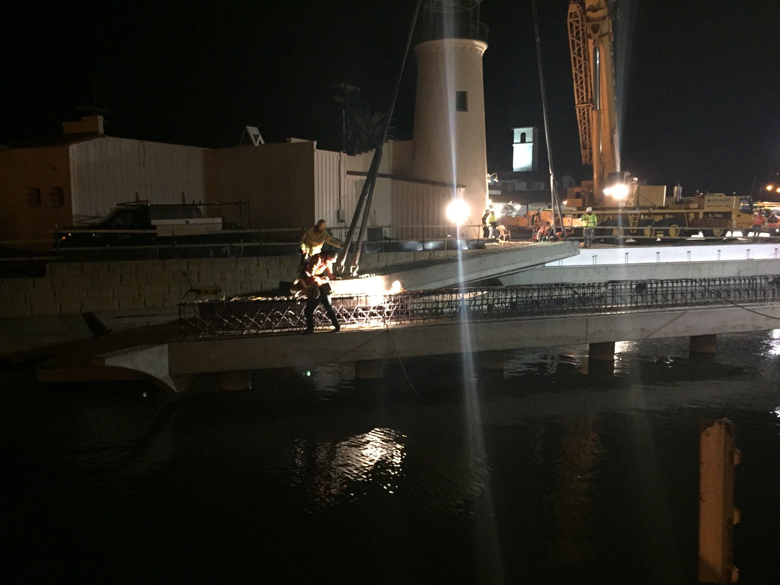 Nighttime construction scene with workers, a crane, and machinery on a building rooftop, illuminated by bright work lights.