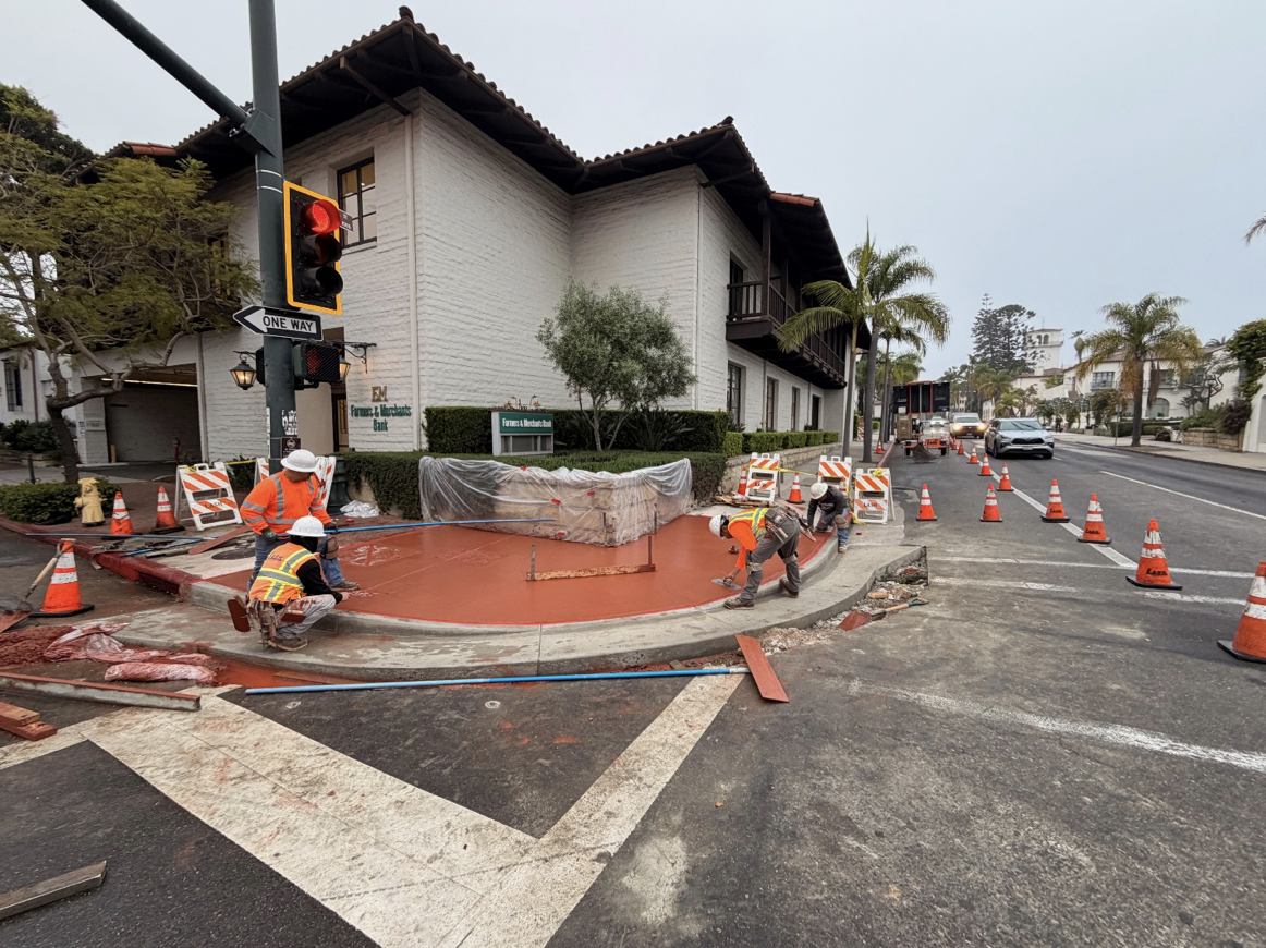Construction workers in safety vests and helmets working on a sidewalk and street corner with traffic cones and barriers in a residential area near a white building and palm trees.