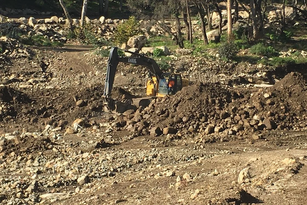 A yellow excavator working in a rocky construction site surrounded by trees.