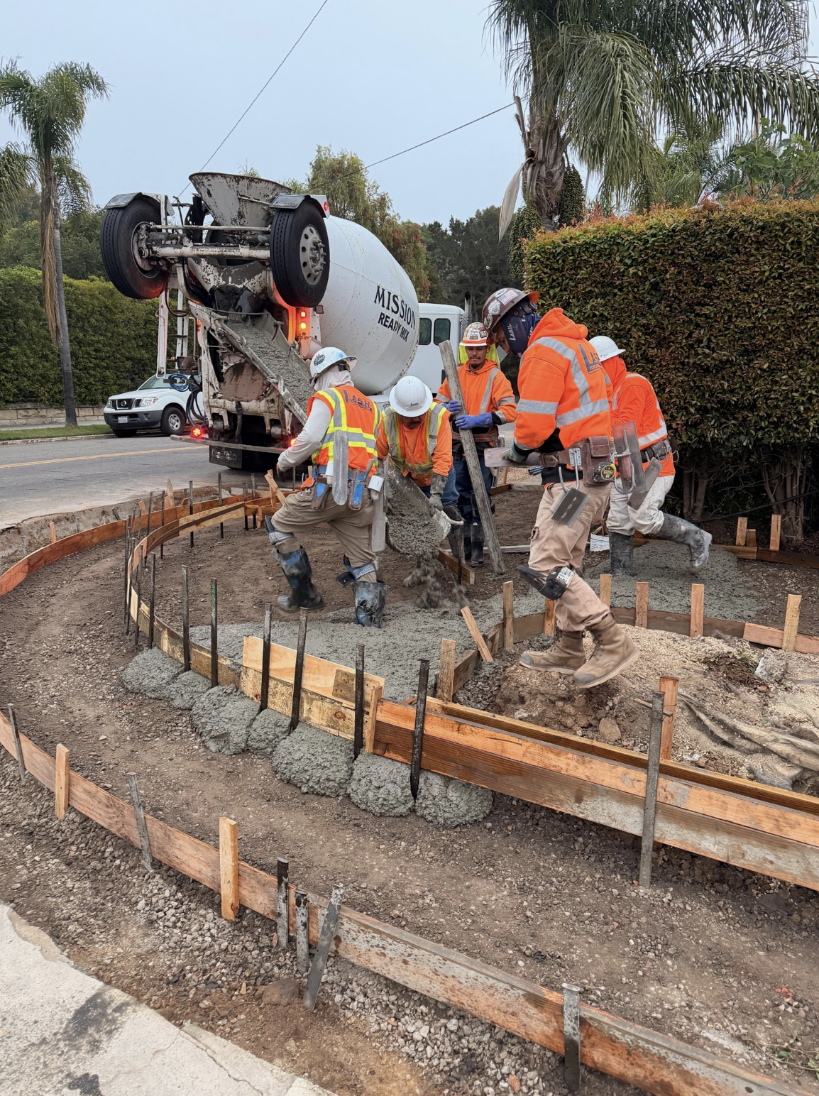 Construction workers are pouring concrete for a curved sidewalk or pathway along a street, with a concrete mixer truck in the background.