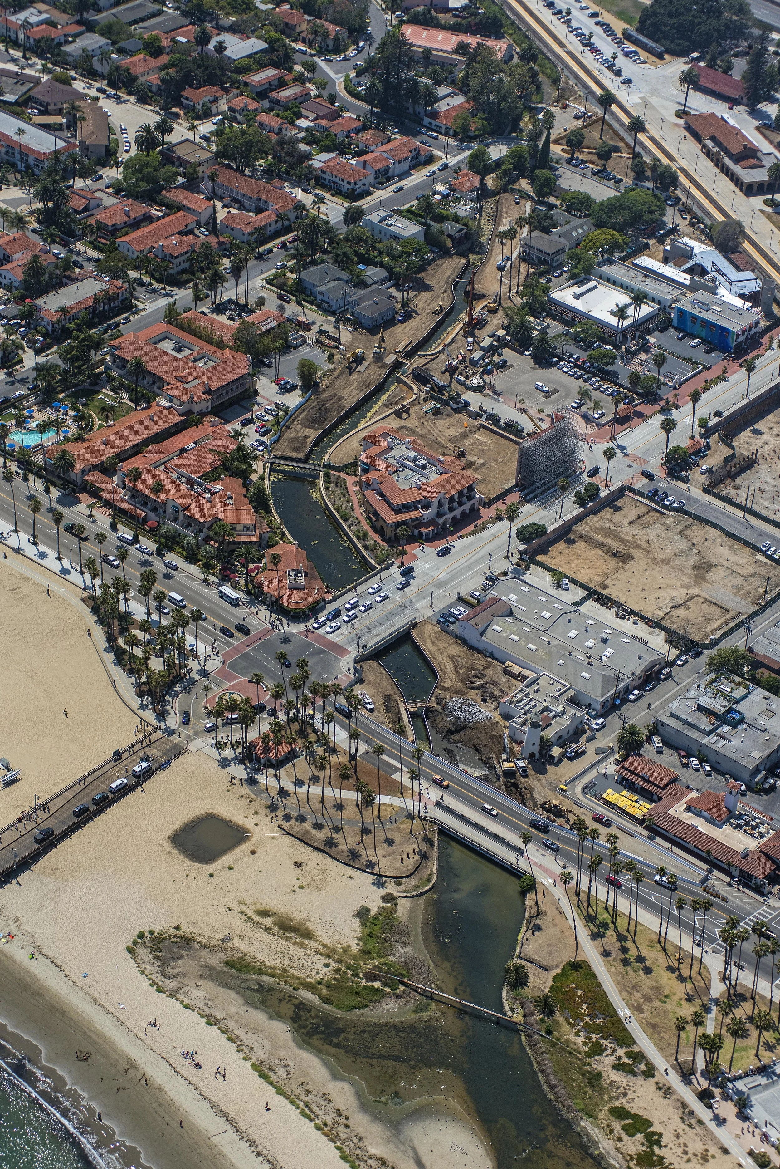 An aerial view of a coastal area featuring a beach with people, a small pond or lagoon, a stream, and numerous buildings including houses, commercial structures, and construction sites, with trees and parking lots.