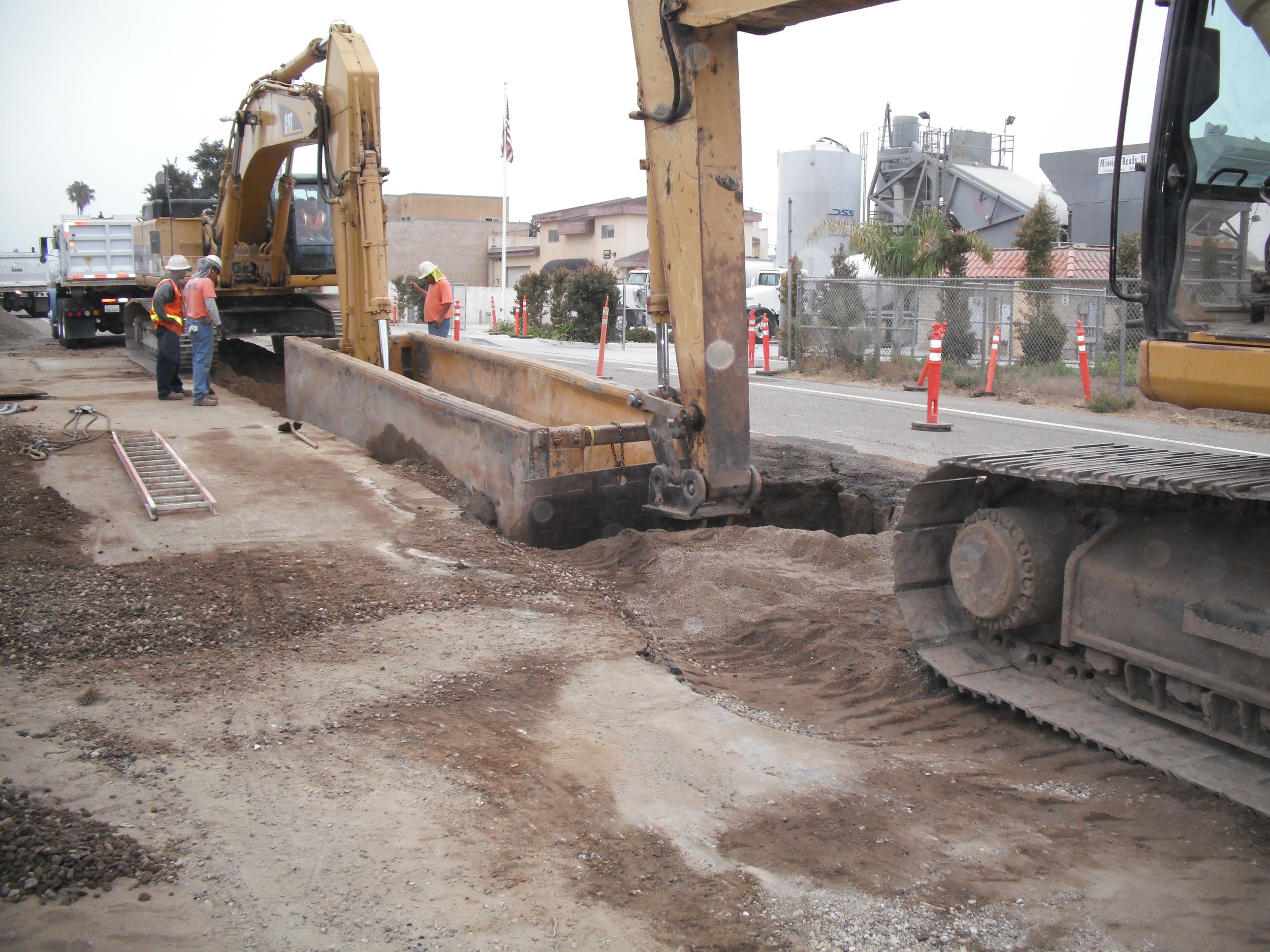 Construction workers are working on a road repair project with heavy machinery and equipment, surrounded by orange safety cones and a chain-link fence.