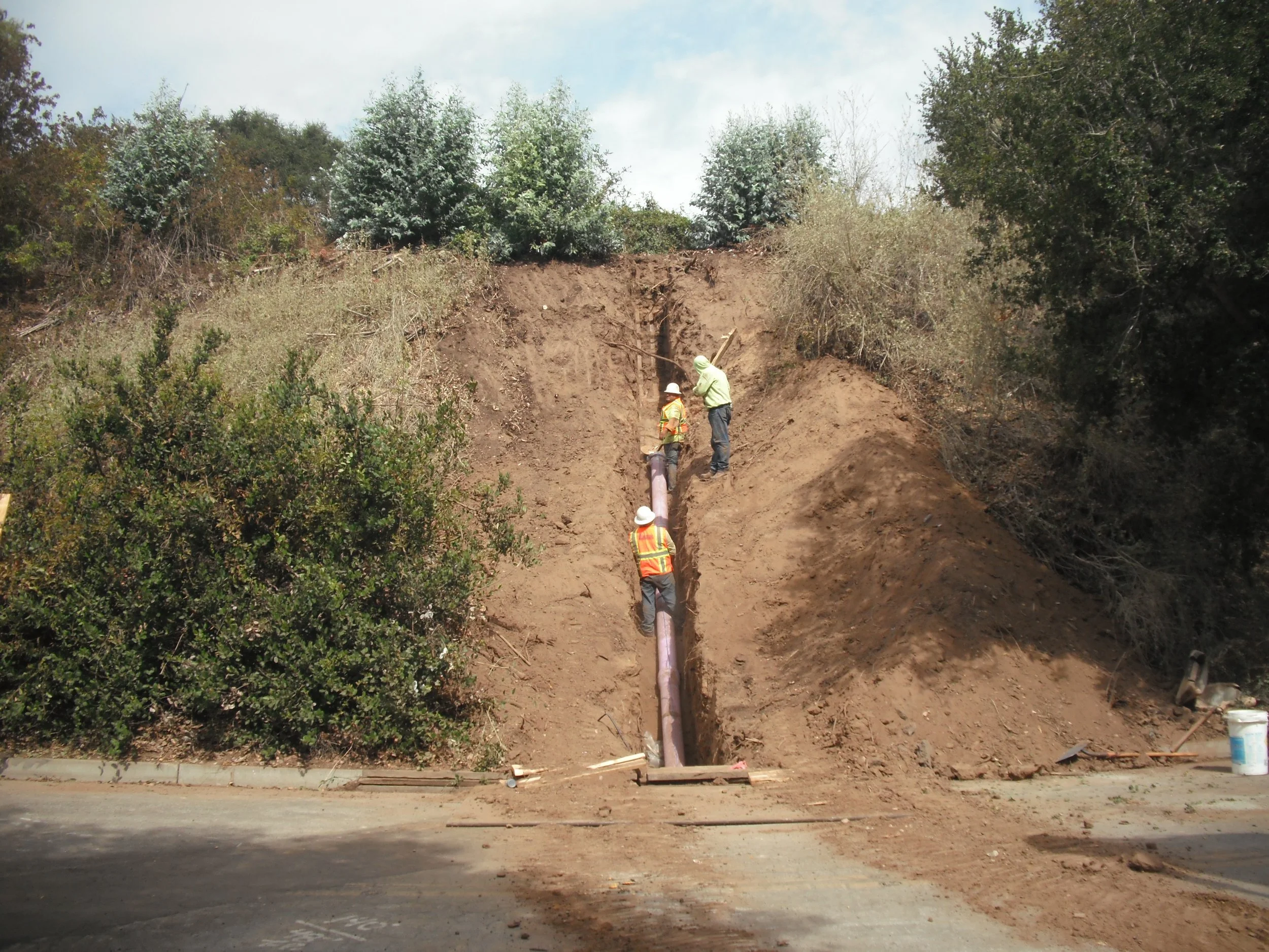 Workers installing underground pipeline on a hillside with trees in the background.