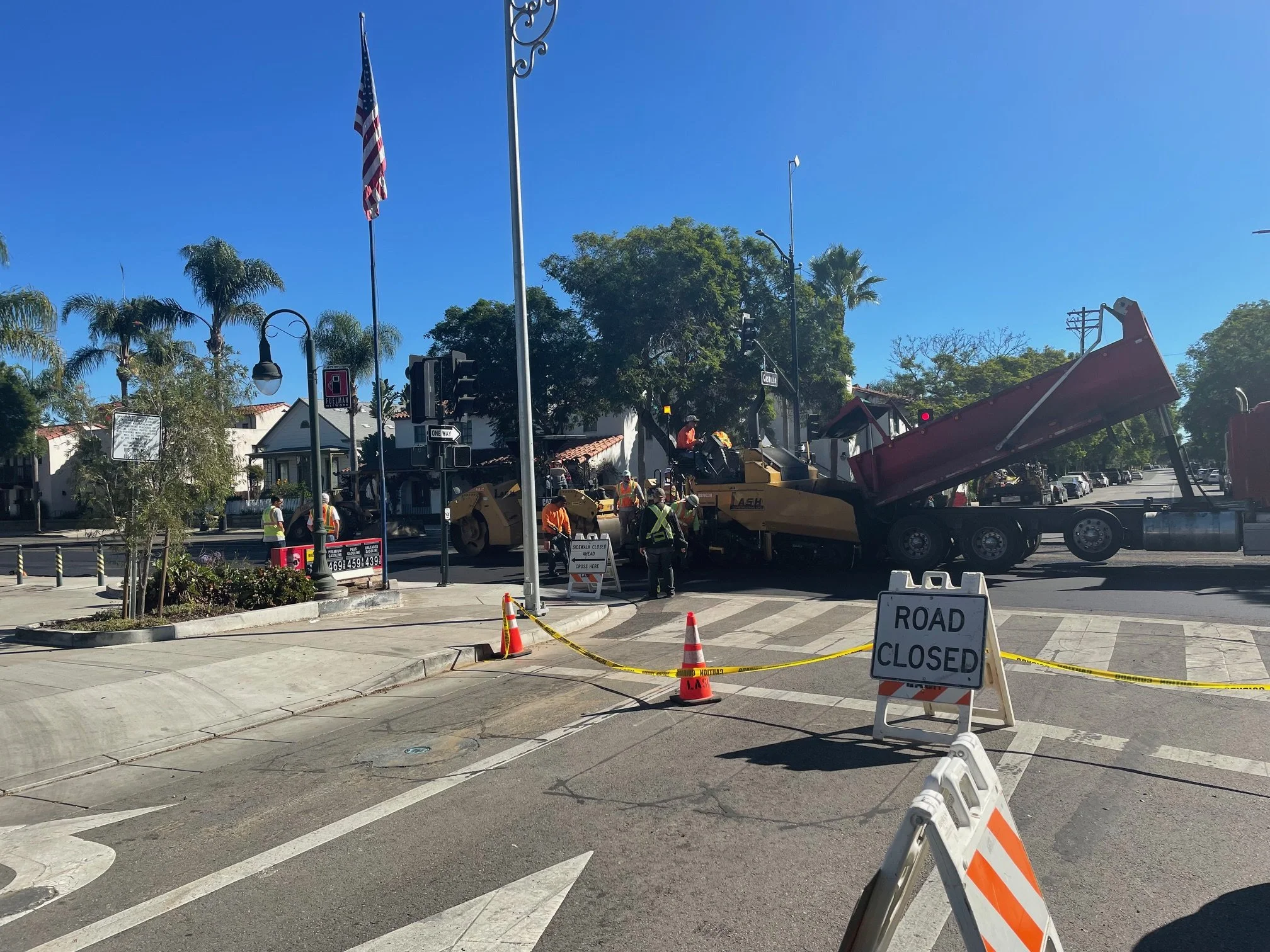 Street construction site with workers, heavy machinery, and signs indicating road closure.