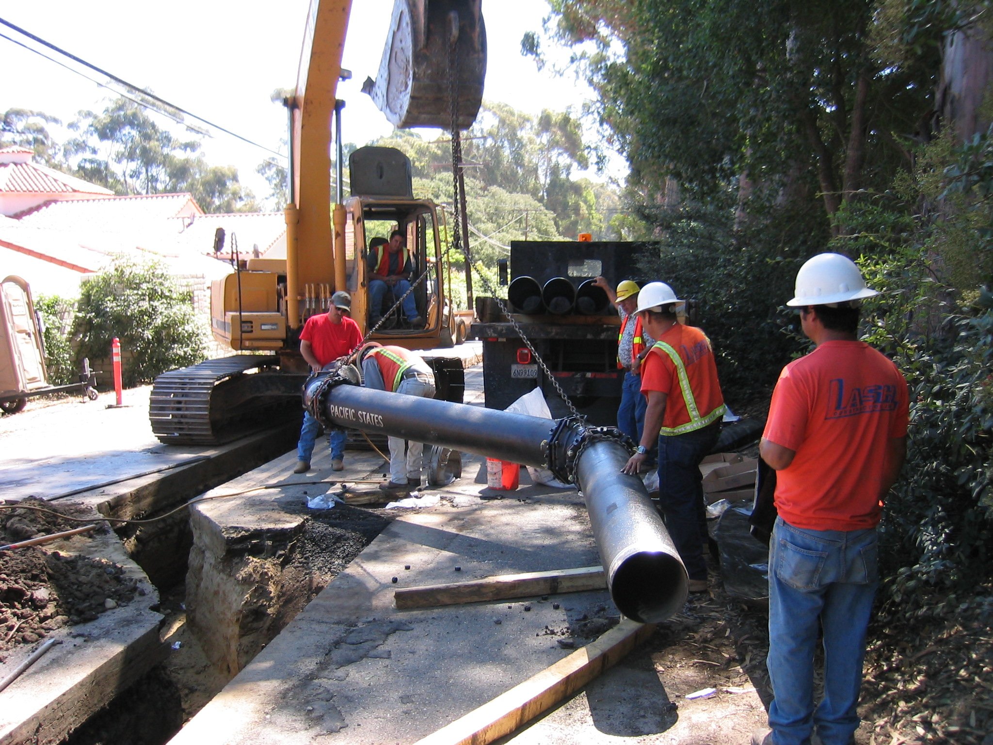 Workers in hard hats and safety vests working on underground pipe installation or repair at a construction site, with excavated trench and equipment.