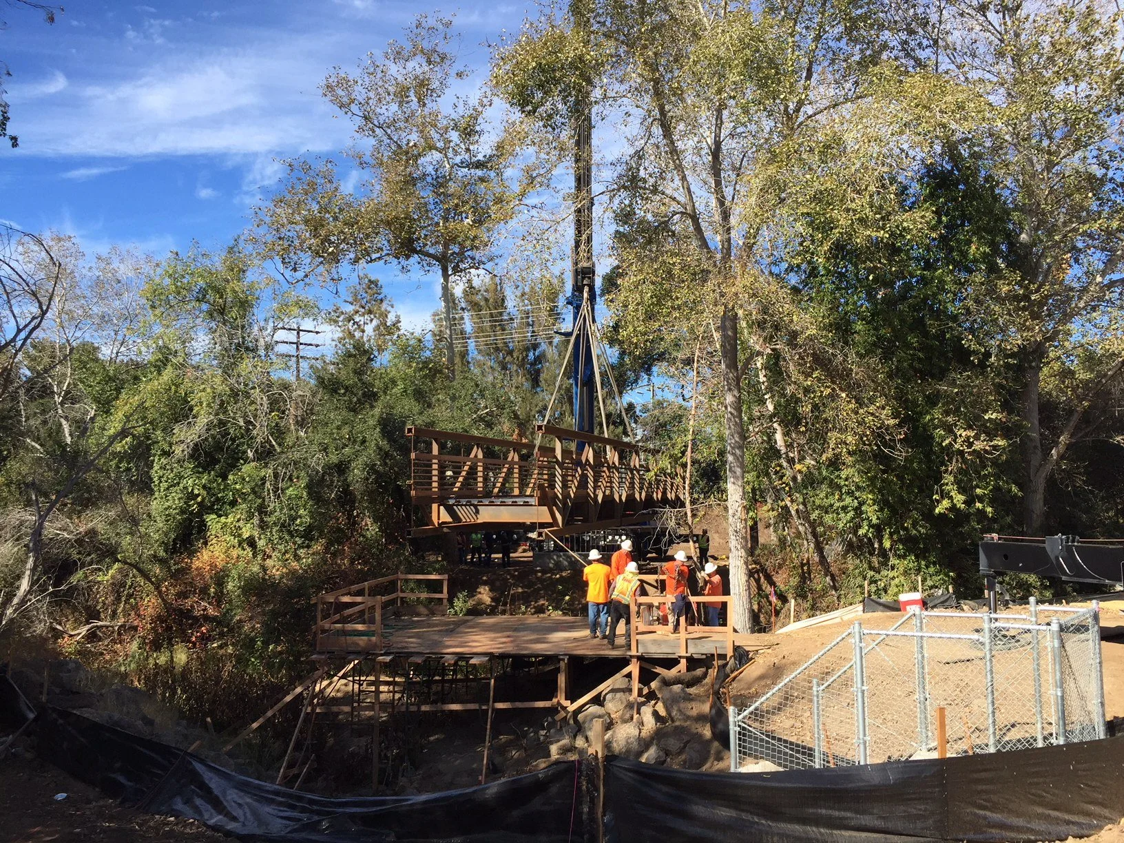 Construction workers assembling a large bridge structure in a wooded area with trees and blue sky in the background.