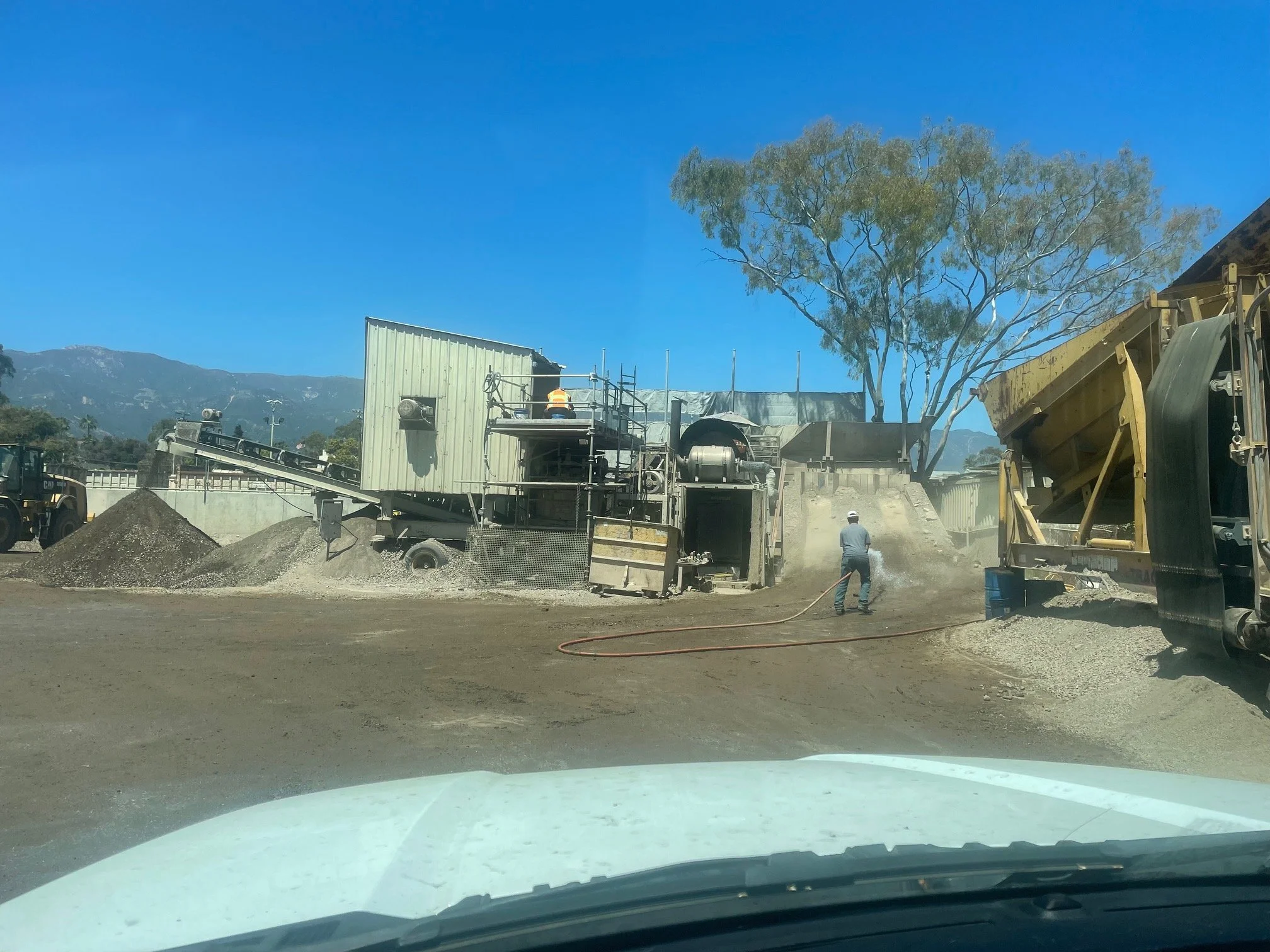 Construction site with workers operating machinery, a large tree, and mountains in the background under a clear blue sky.