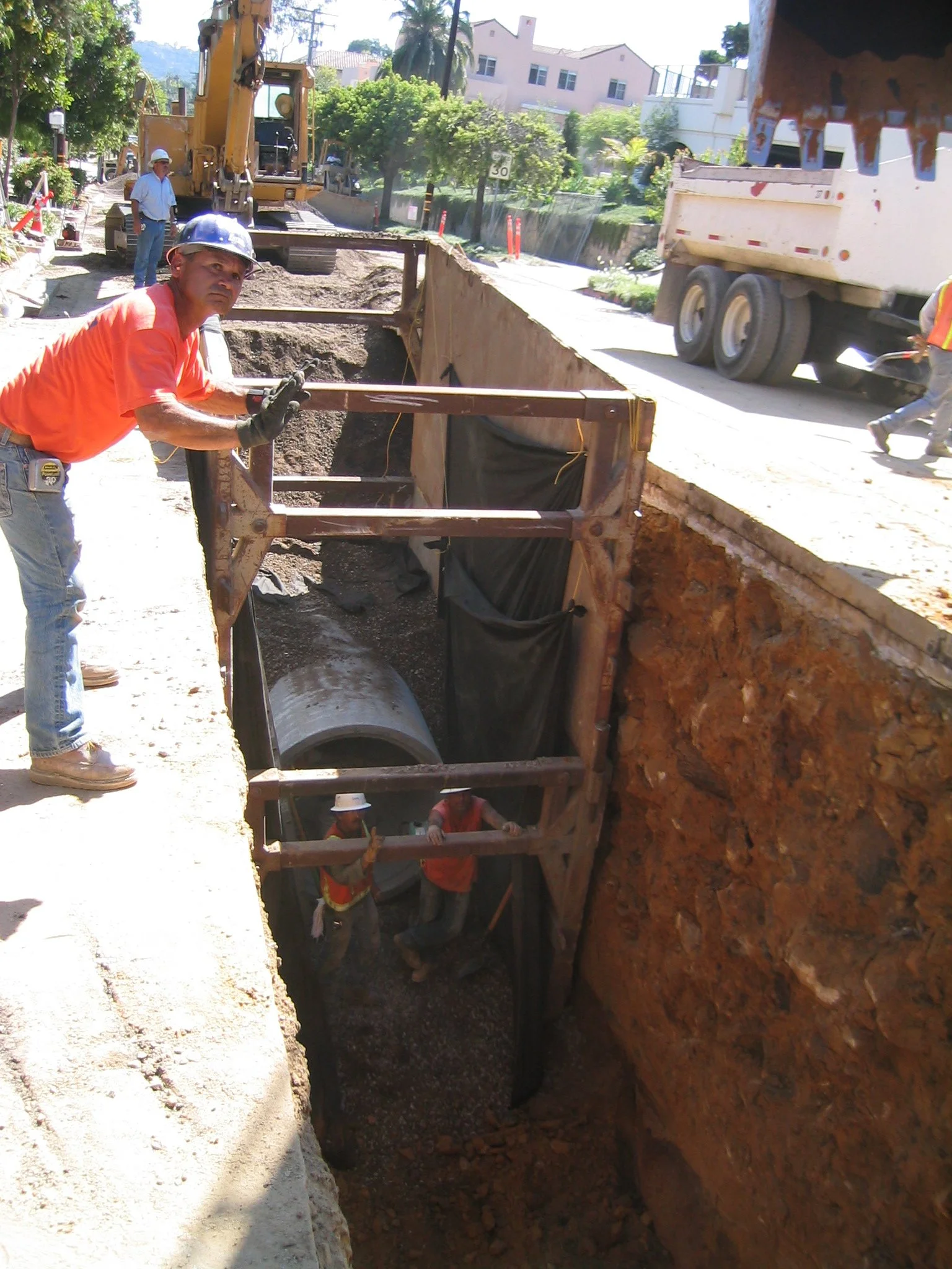 Construction workers installing underground pipes on a street, with a backhoe and truck nearby, in a residential neighborhood.