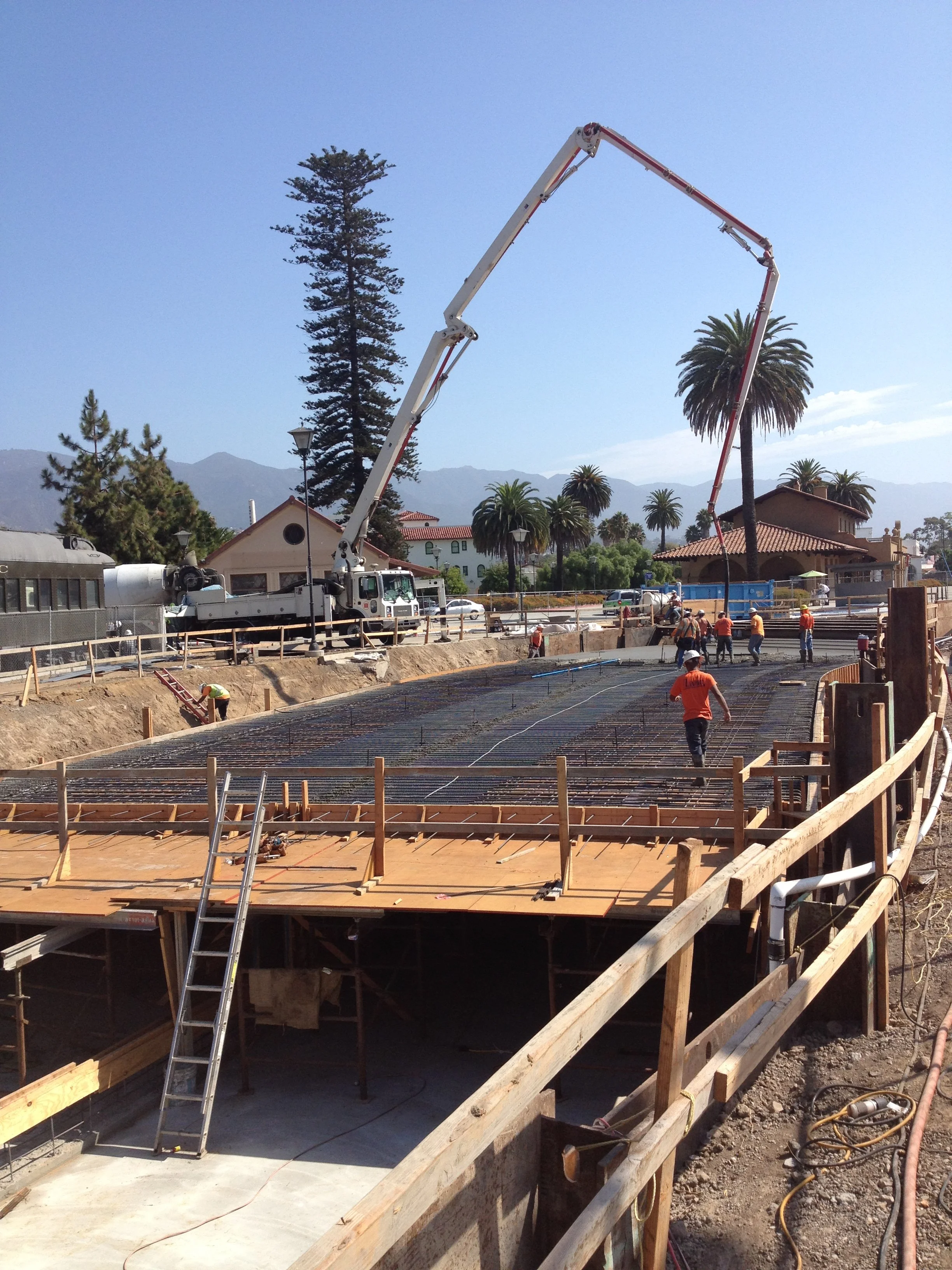 Construction site with workers pouring concrete, wooden framework, and a large crane in background under a clear sky with mountains and palm trees.