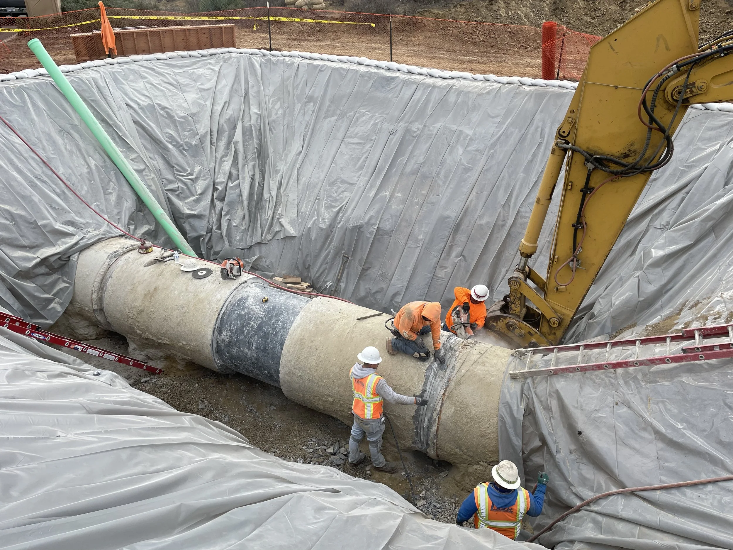 Construction workers in safety vests and helmets work on a large pipeline in a deep excavation lined with plastic sheeting, with construction equipment and safety fencing around the site.