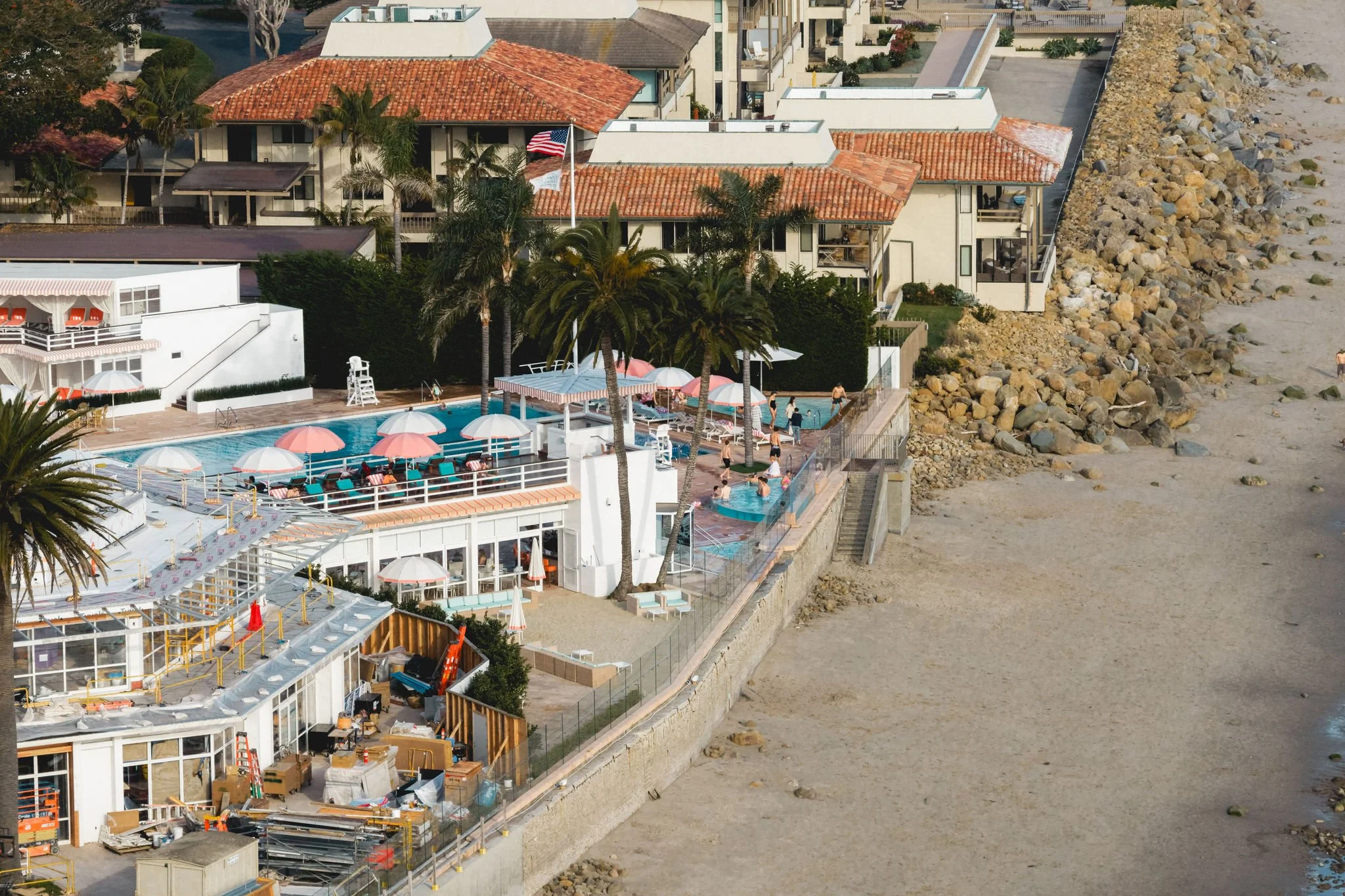 An aerial view of a beachside hotel with a swimming pool, surrounded by palm trees, outdoor seating, and buildings with red-tile roofs. The beach and rocky shoreline are on the right side of the image.