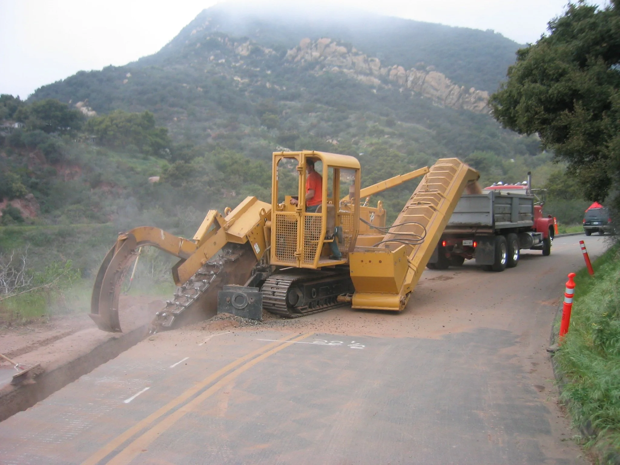 A yellow bulldozer working on road construction, moving dirt, with hills and trees in the background.