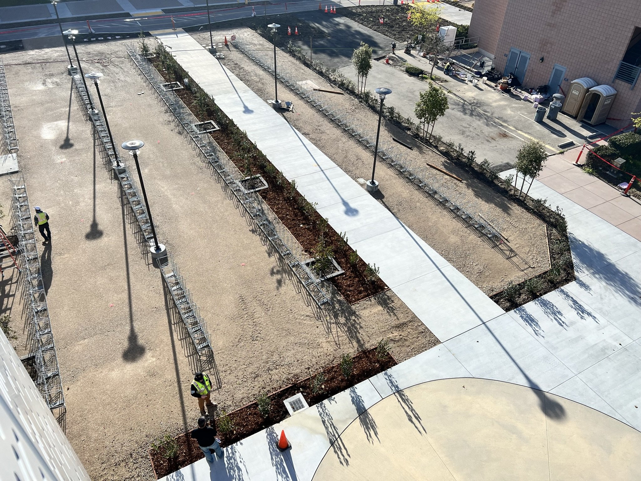 Construction site with workers and fencing, trees along the sidewalk, and parking lot in the background.