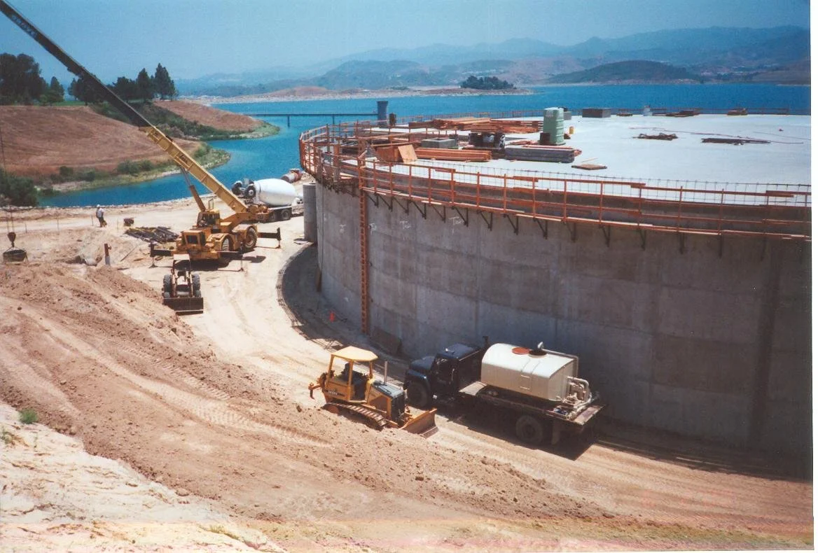 Construction workers and heavy machinery building a large concrete dam near a body of water, with hills and mountains in the background.