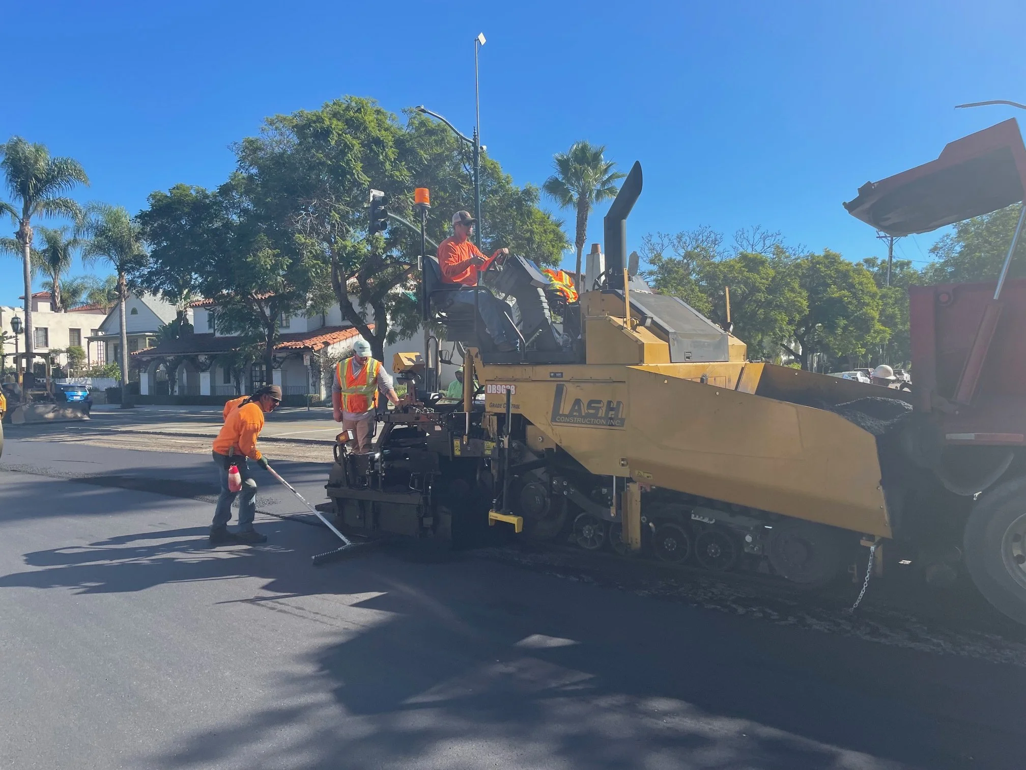 Workers paving a road with heavy machinery and safety vests under a bright blue sky, with trees and buildings in the background.