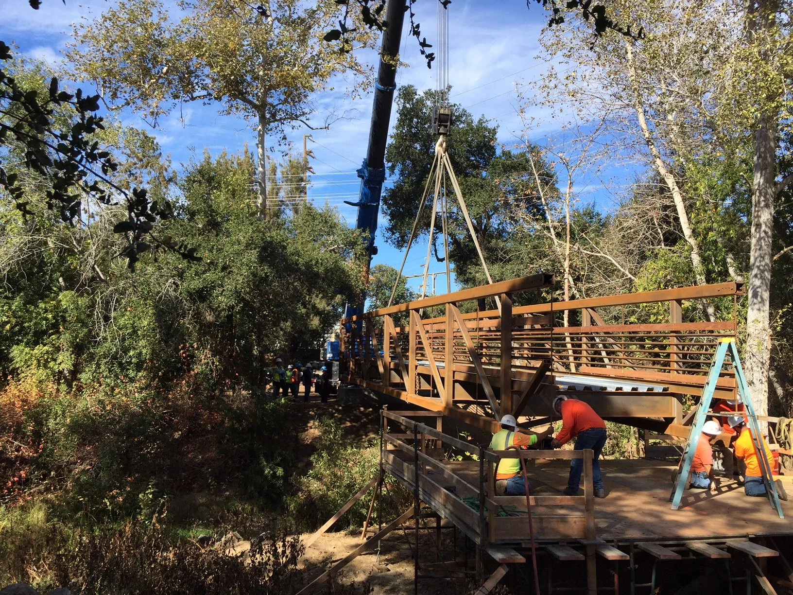 Construction workers building a wooden platform or bridge in a wooded outdoor area with trees and a crane lifting materials.