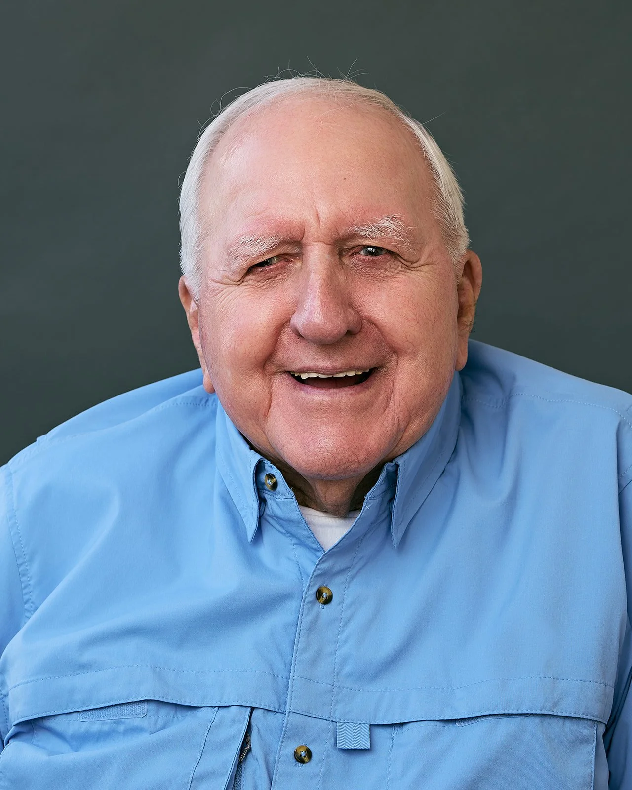 An elderly man with white hair and a smile, wearing a light blue button-up shirt, against a dark background.