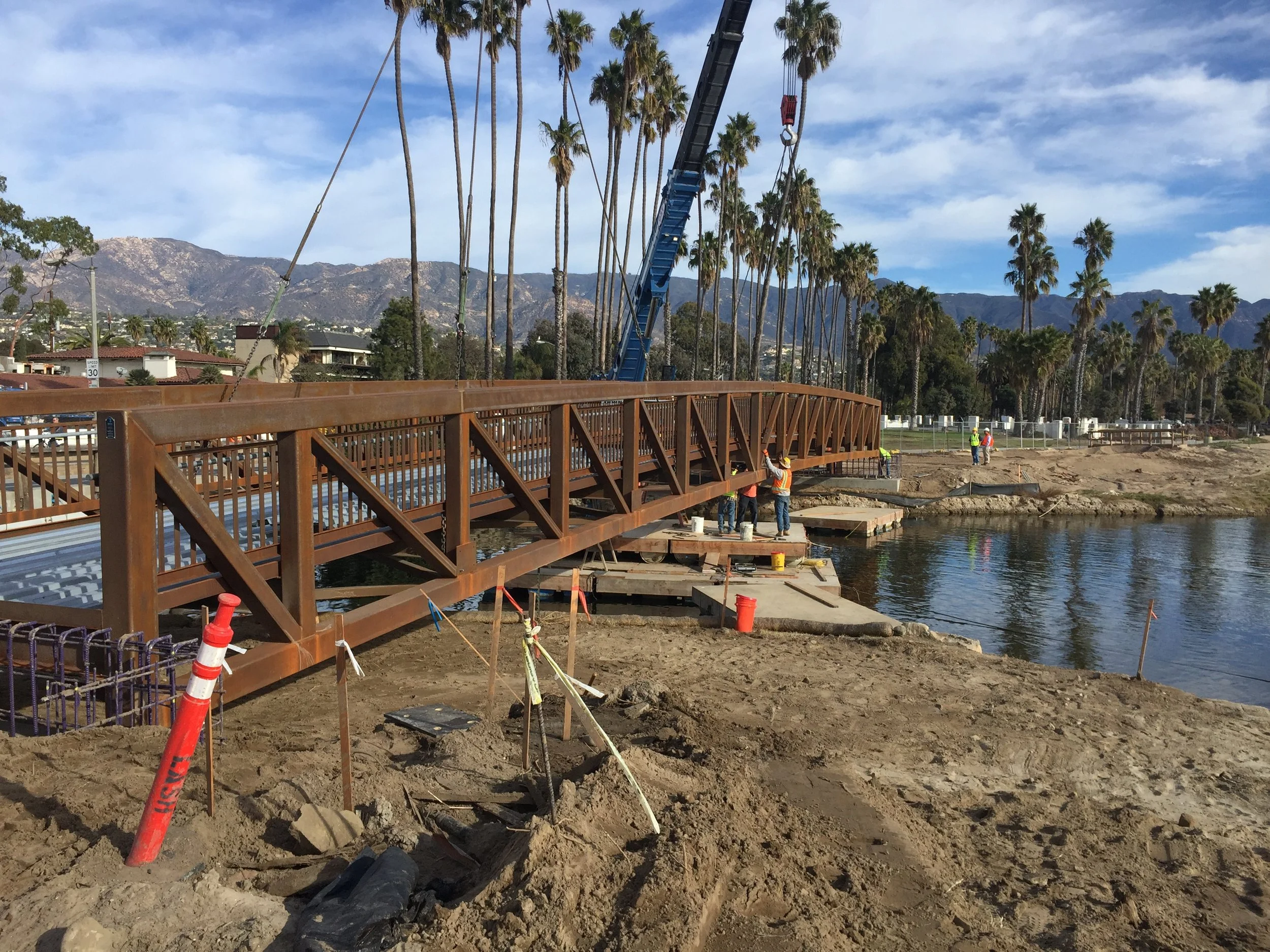 Construction workers building a bridge over water with a scenic background of palm trees and mountains on a clear day.