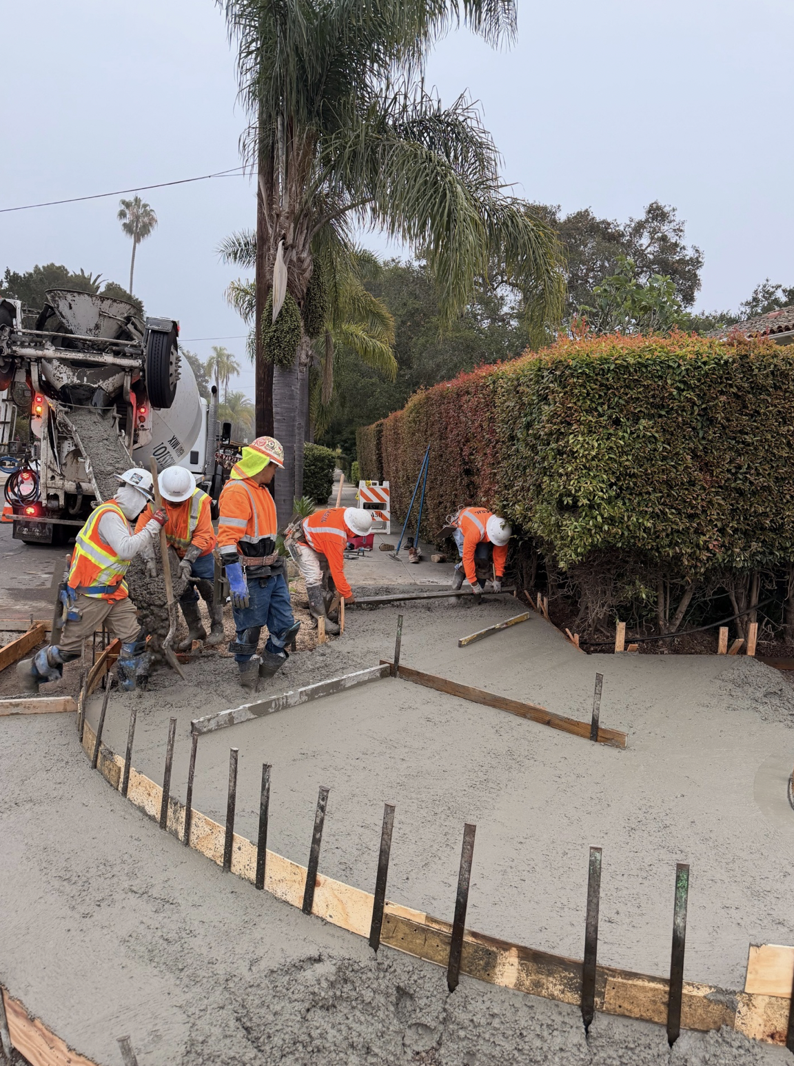 Construction workers in safety gear are pouring and smoothing concrete for a sidewalk or driveway on a residential street, with trees and bushes in the background.