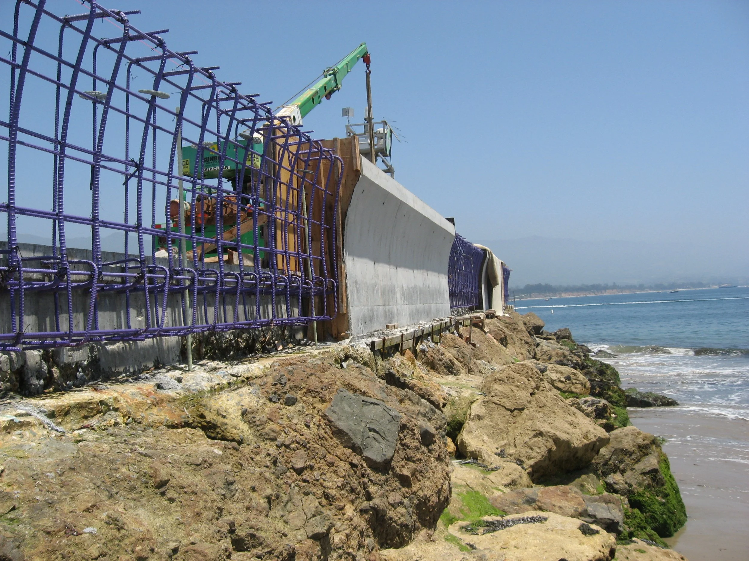 Construction site along a rocky shoreline with a crane and fencing, overlooking the ocean under a clear blue sky.