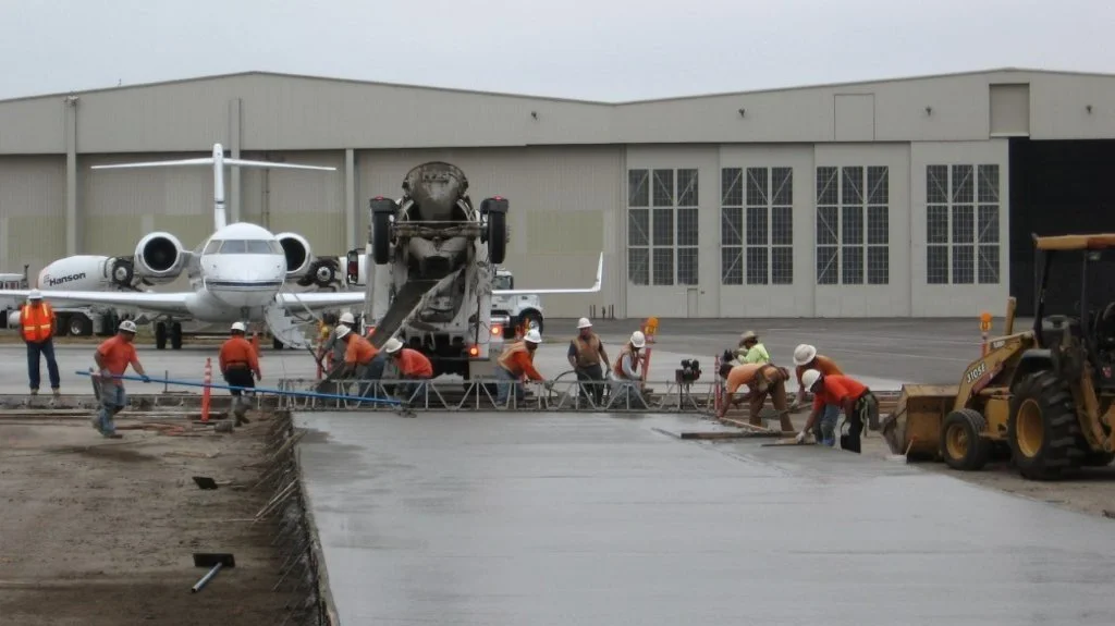 Construction workers pouring and smoothing concrete on a runway at an airport, with an airplane and a hangar in the background.