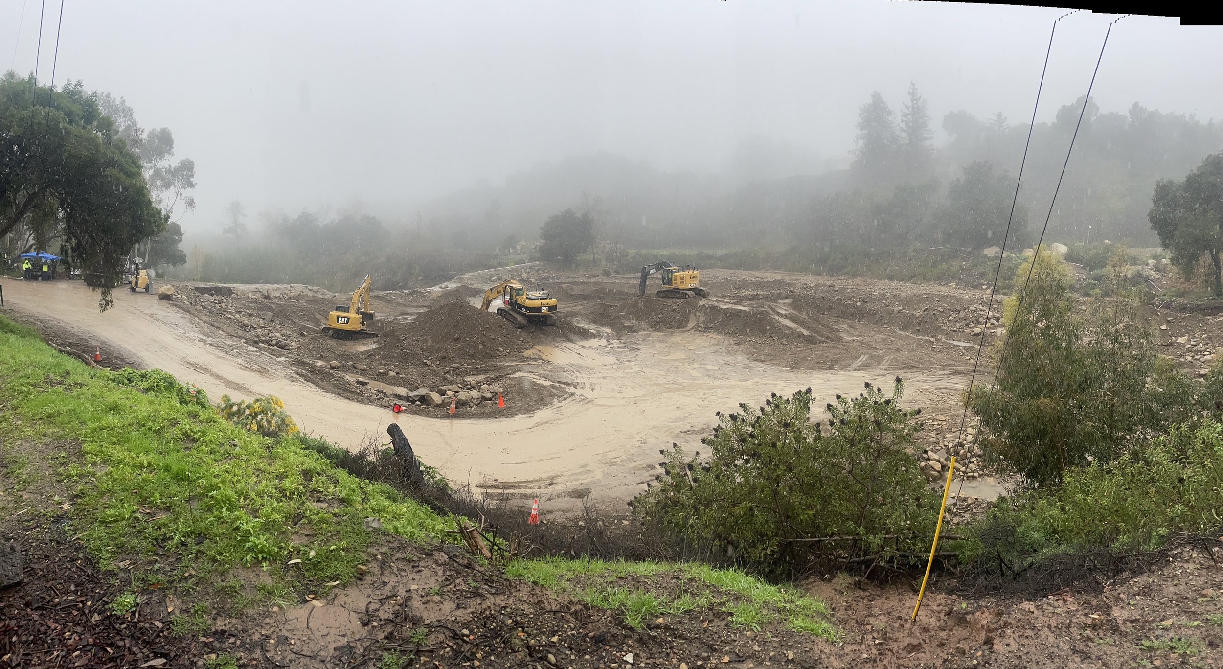 Construction site with three yellow excavators working on a dirt hill in foggy weather, with trees in the background and a muddy road in the foreground.