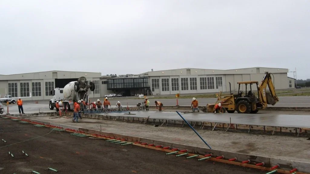 Construction workers pouring concrete on a large outdoor construction site, with a cement mixer and a backhoe, in front of a modern building.