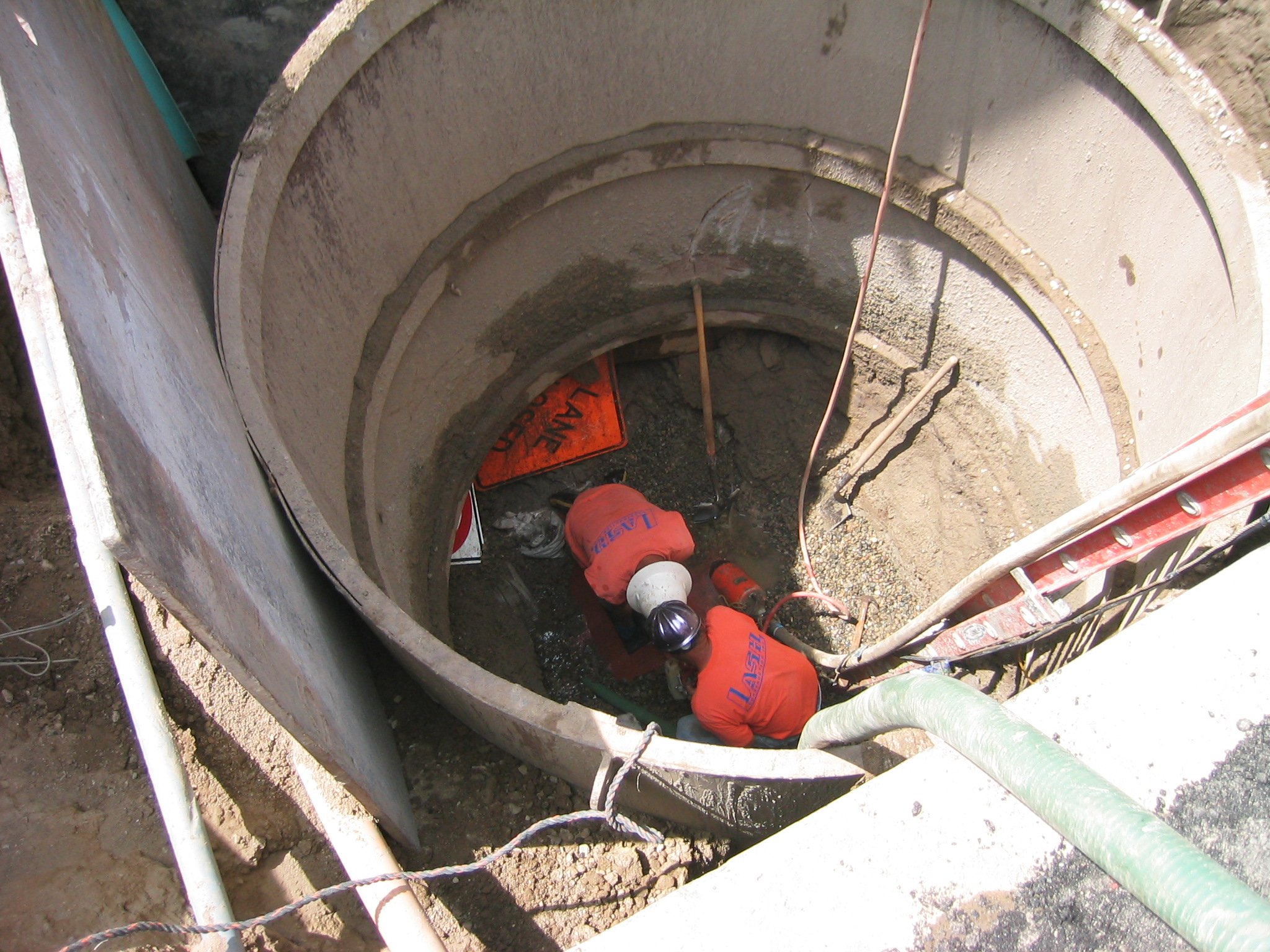 Construction workers inside a deep manhole, working on sewer or drainage pipes, with safety equipment and tools, surrounded by concrete walls.