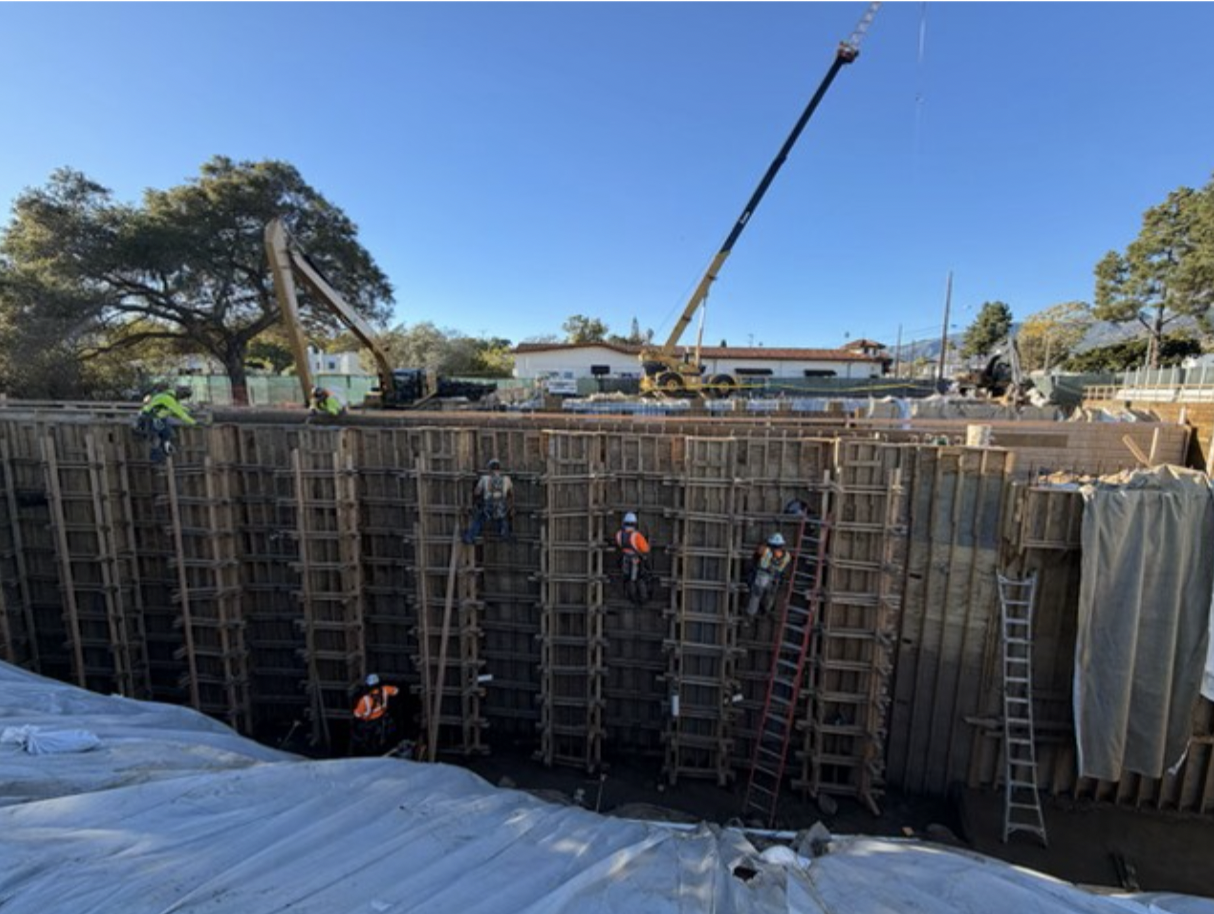 Construction workers building a deep foundation with wooden formwork, using ladders and a crane on a construction site.
