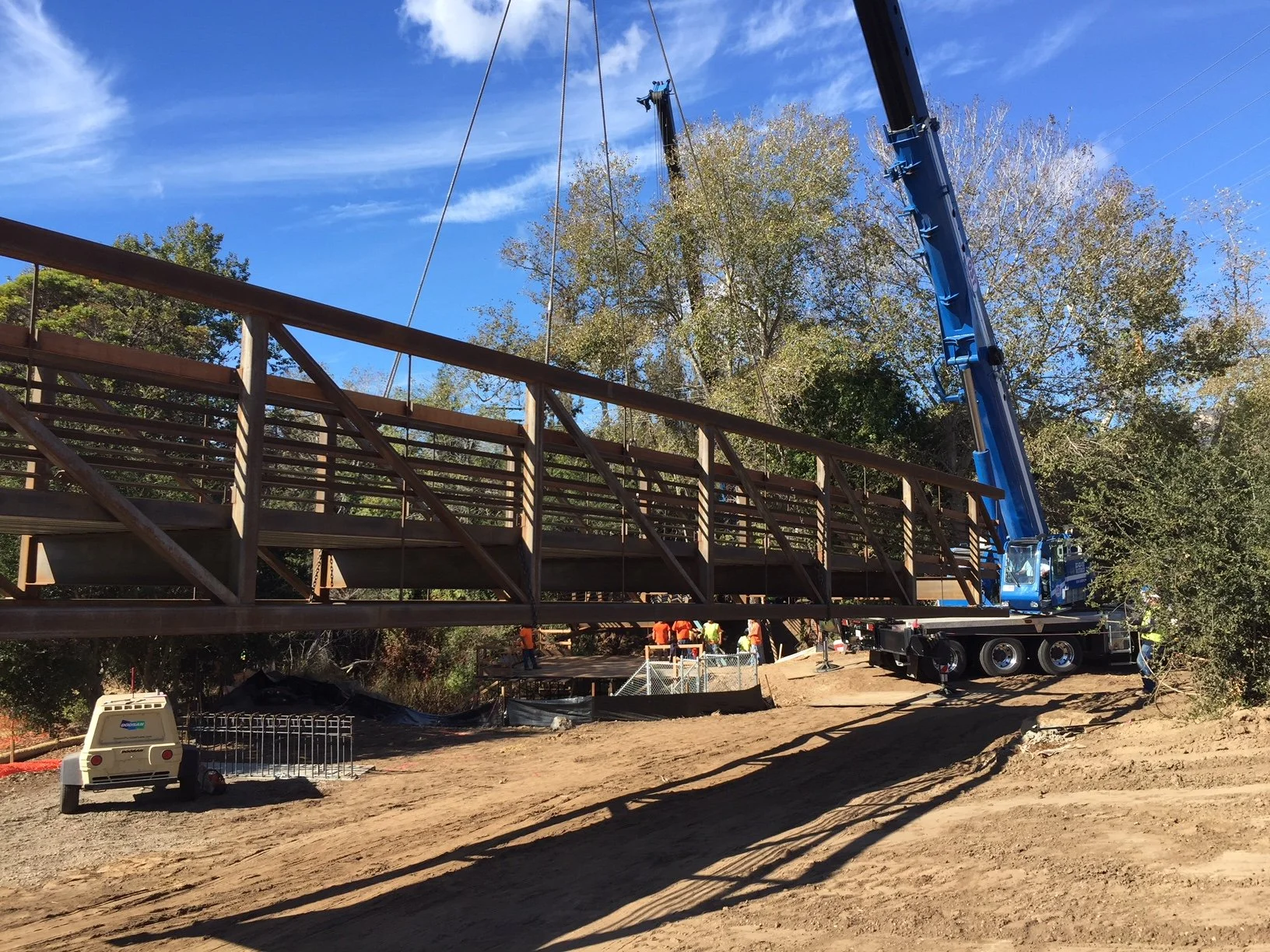 Construction workers building a new bridge with a crane lifting the bridge structure into place, set against a background of trees and a blue sky.
