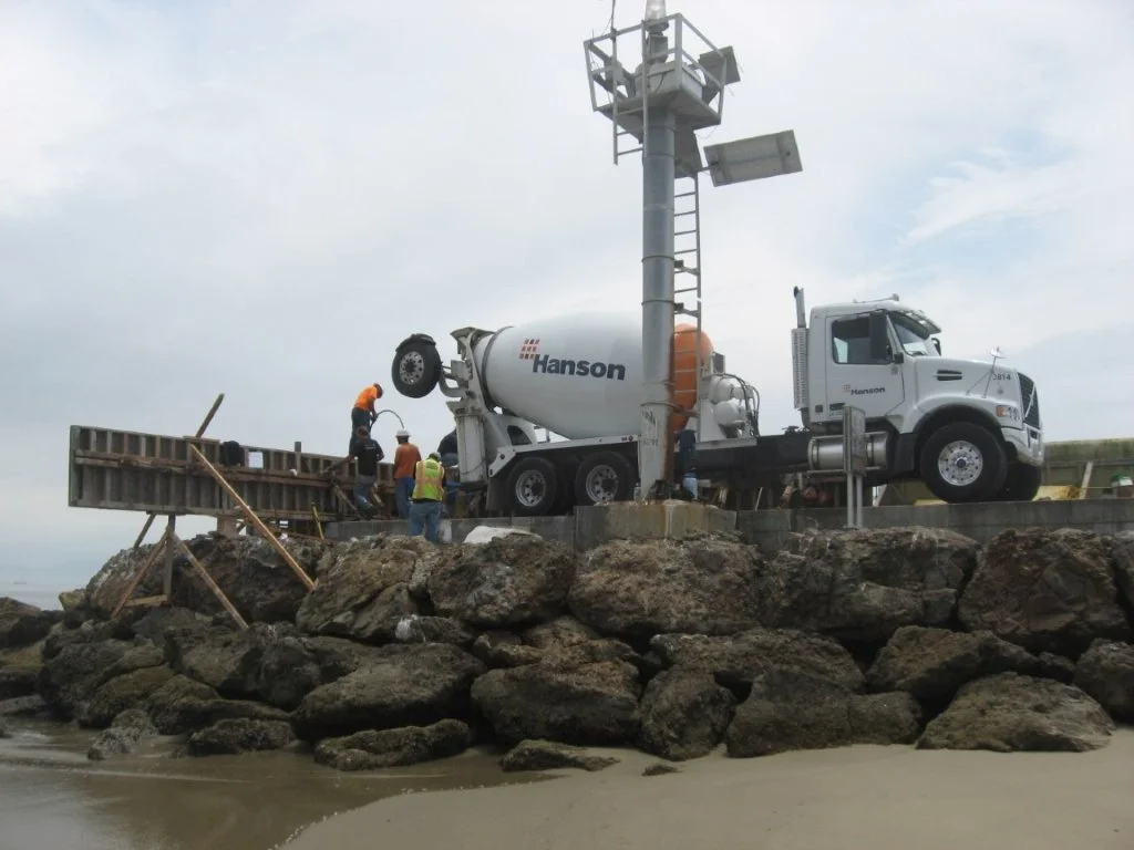 Construction workers guide a cement mixer truck on a rocky shoreline with a storm drain pole.