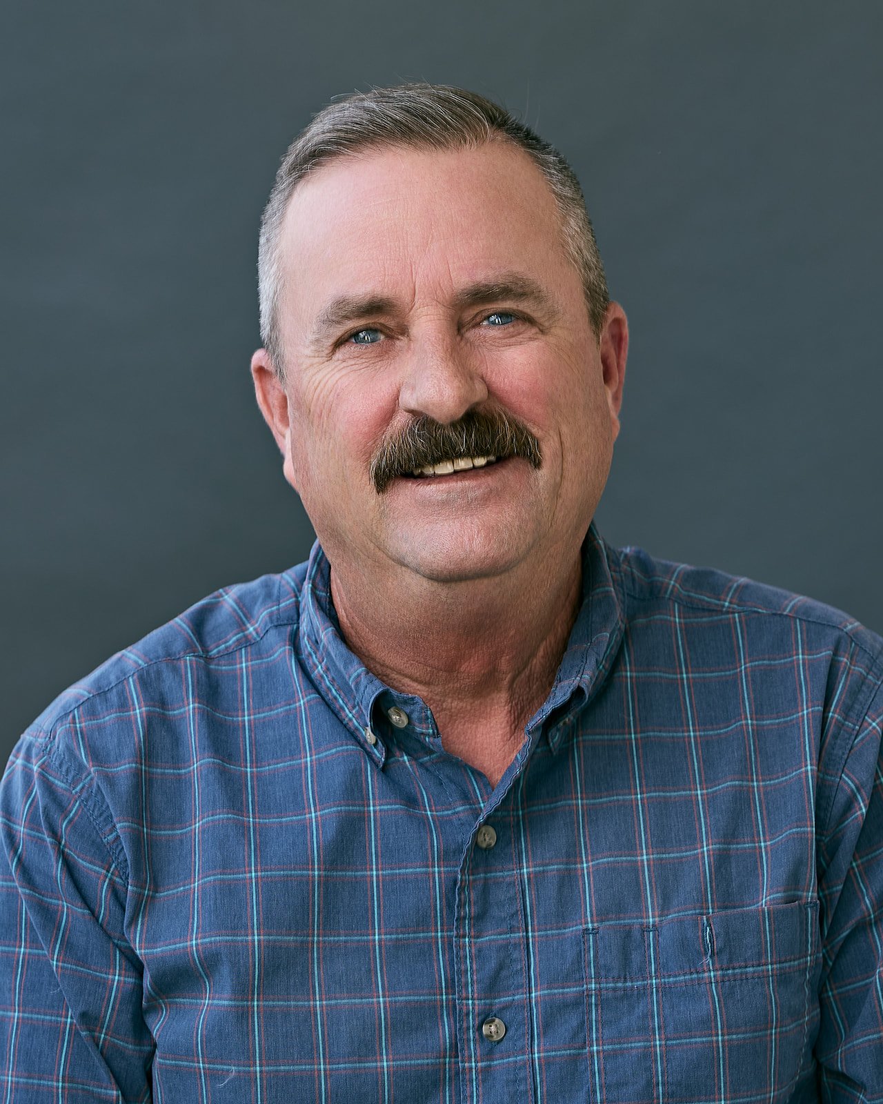 A middle-aged man with short gray hair, a thick mustache, and blue eyes, smiling and wearing a blue plaid shirt, against a dark gray background.