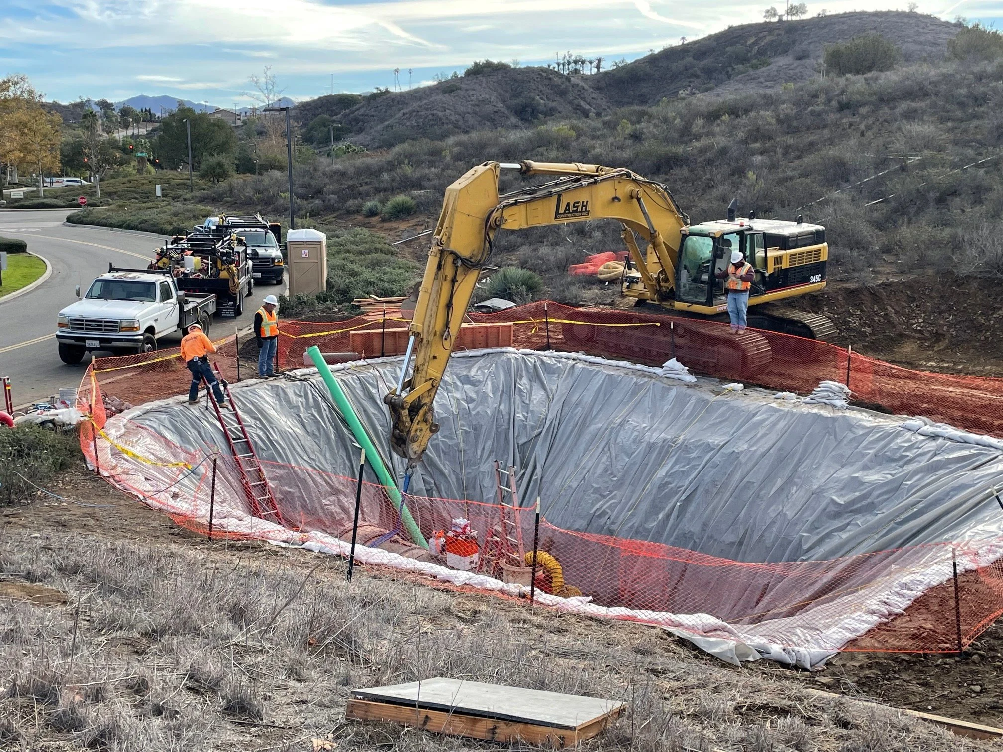 Construction site with an excavator digging a large hole, surrounded by safety fencing and construction workers, near a road with hilly terrain in the background.