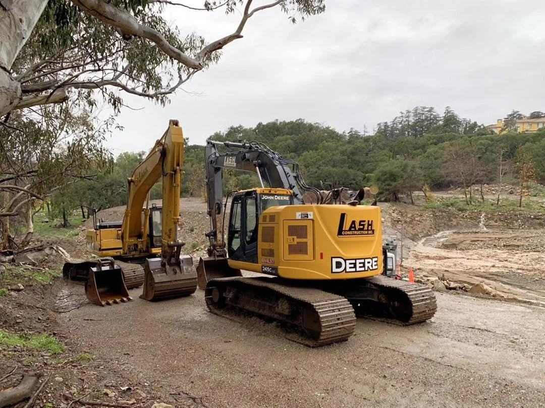 A John Deere construction excavator working on a dirt road in a hilly area, with trees and cloudy sky in the background.