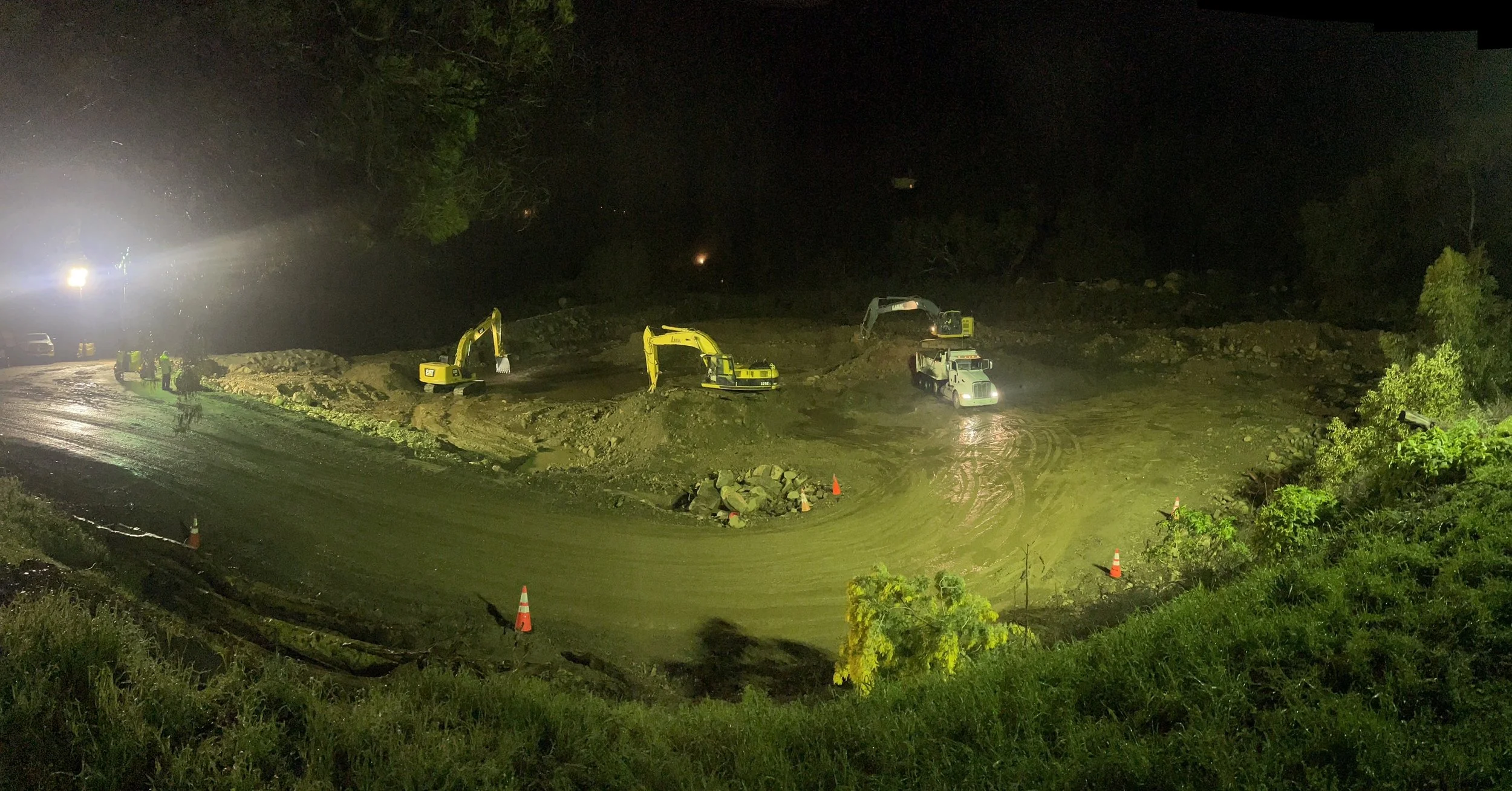 Nighttime construction site with excavators, a dump truck, safety cones, and workers, and military vehicles with bright lights illuminating the area.