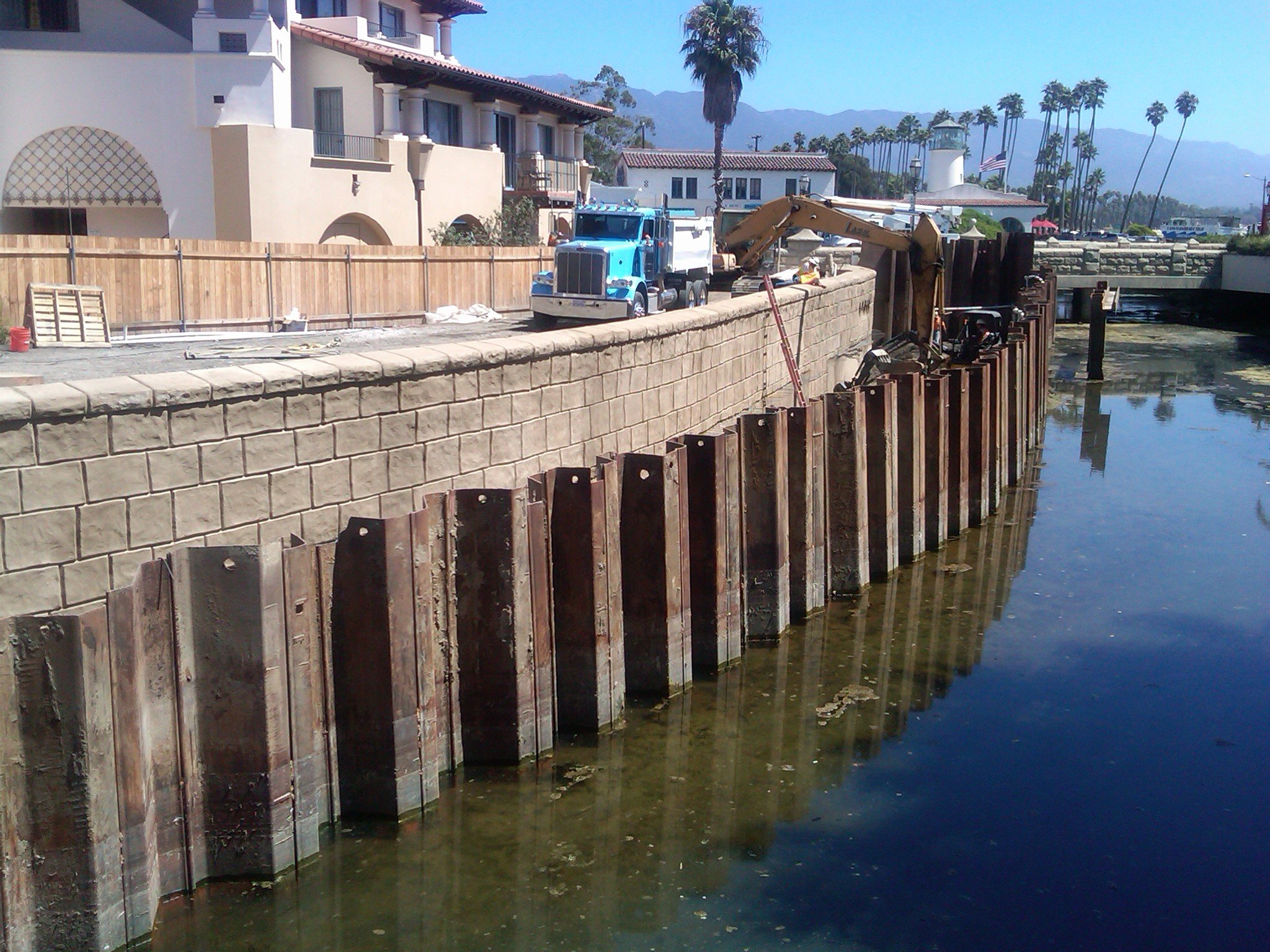 A construction site along a canal or waterway, with a stone wall and wooden support beams, a yellow excavator, a blue truck, and a white building with a red-tiled roof in the background. Tall palm trees and mountains are visible in the distance under
