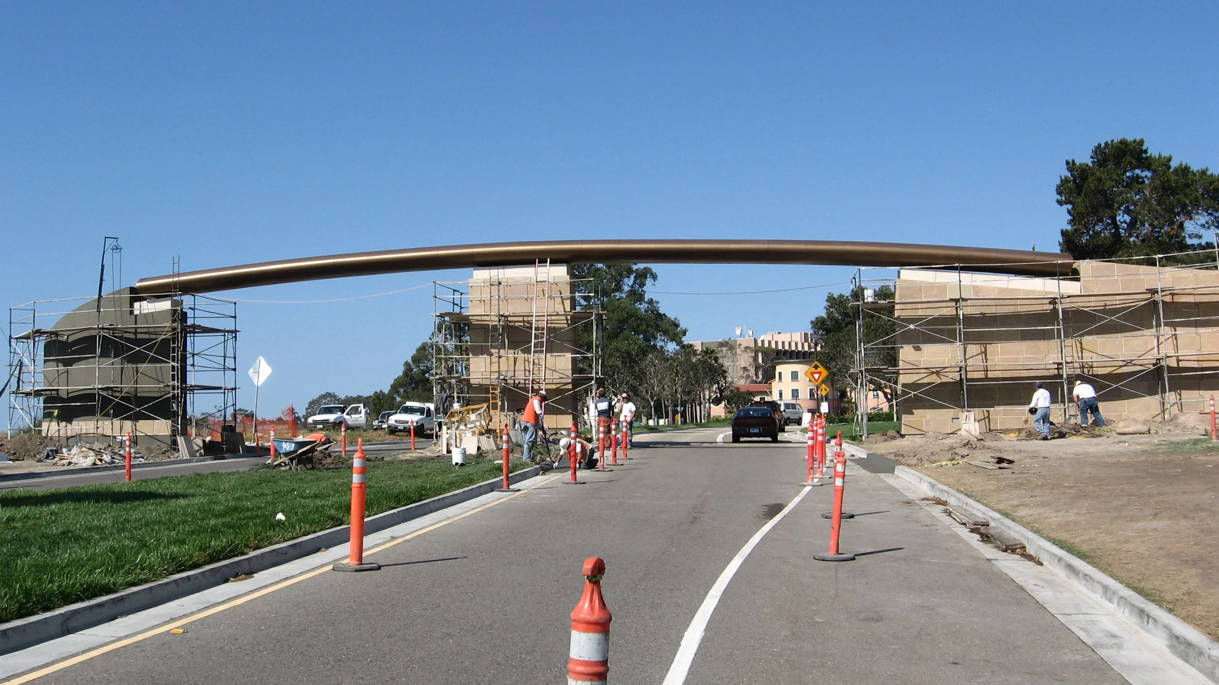 Construction workers installing a large metal arch over a roadway, with scaffolding and orange safety cones around the site, and trees and buildings in the background.