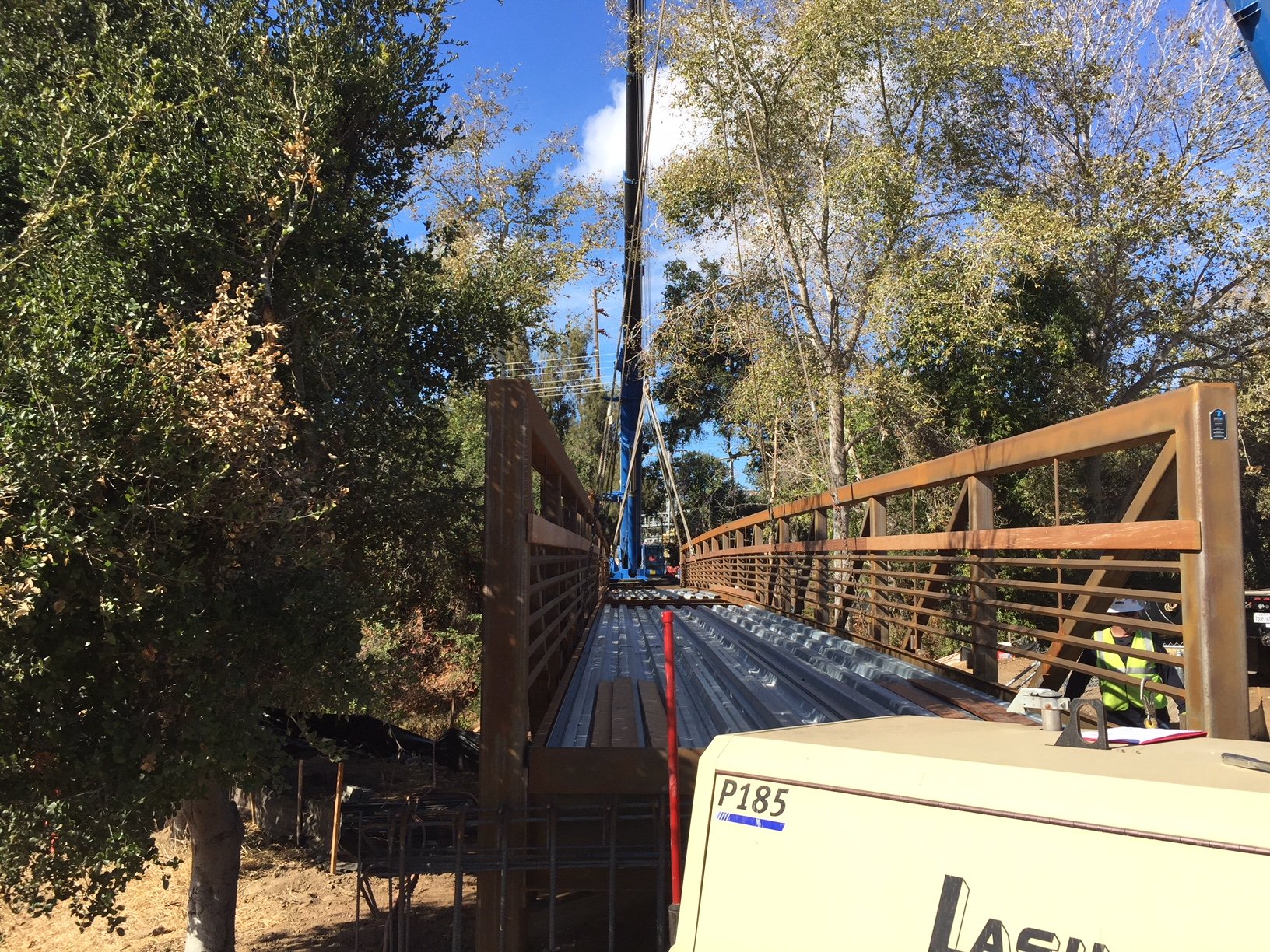 Construction site with a bridge or walkway under construction in a wooded area, with equipment and workers visible.