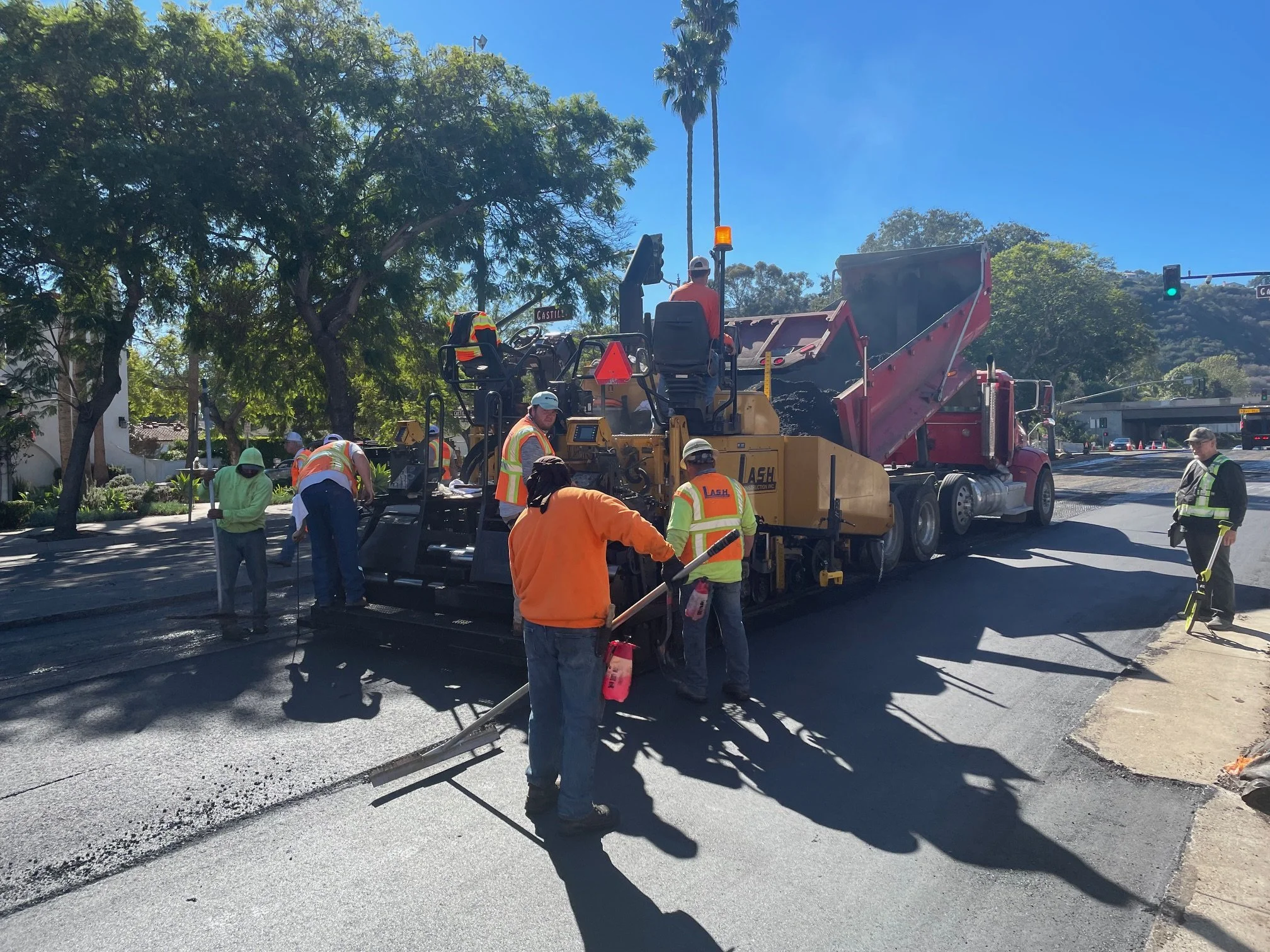 Workers in orange safety vests and hard hats are paving and repaving a street with asphalt. Construction equipment is in use, and some workers are using tools, with traffic lights and trees in the background.