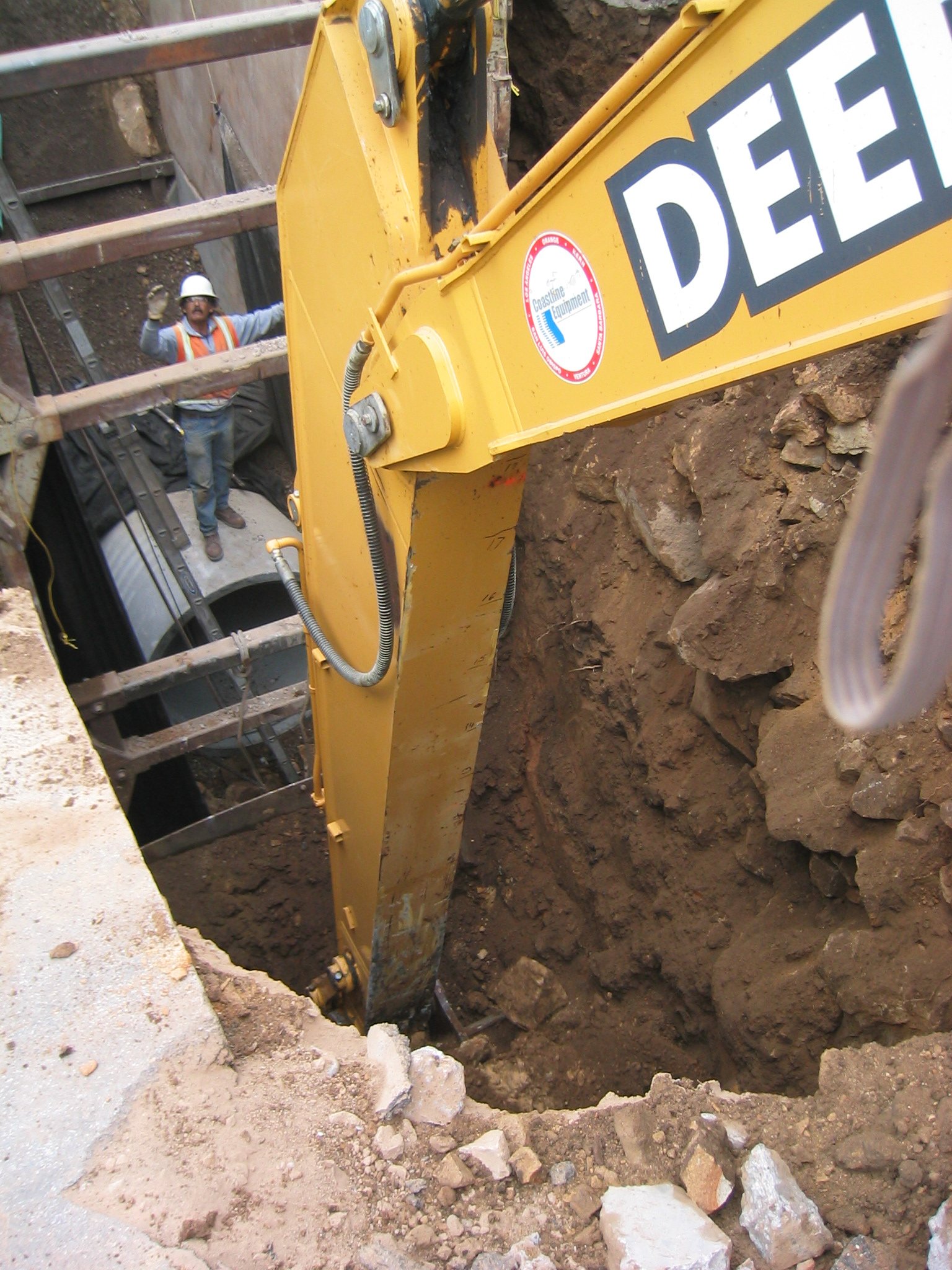 Construction worker with hard hat and safety vest standing in a trench, operating a yellow backhoe digging into the ground.