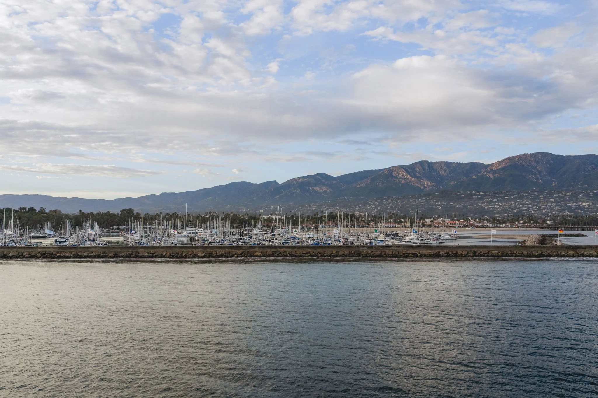 A marina with sailboats docked, a city in the distance, mountains, and a partly cloudy sky.