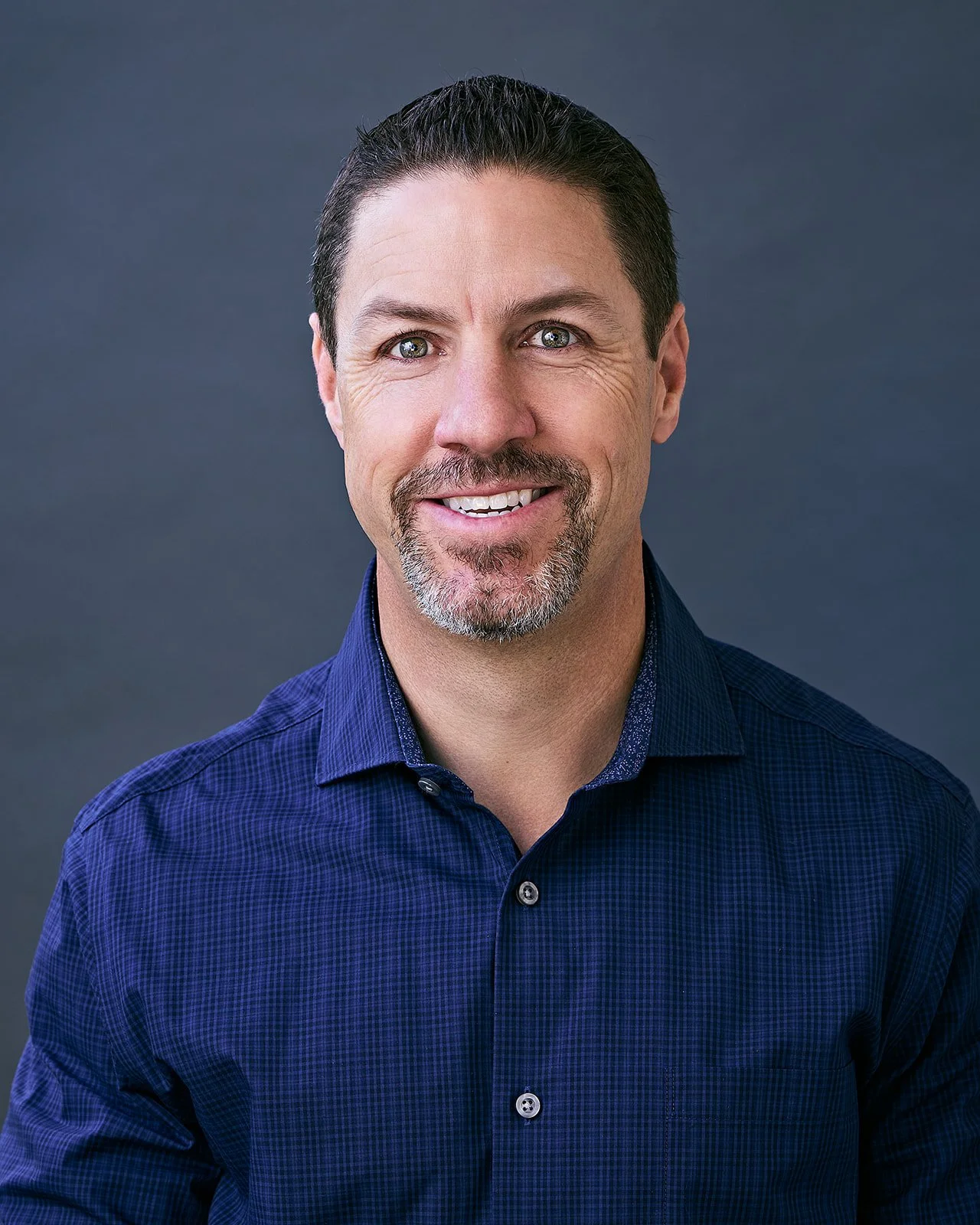Portrait of a man with dark hair, blue eyes, and a goatee, wearing a dark blue checkered shirt, smiling against a gray background.