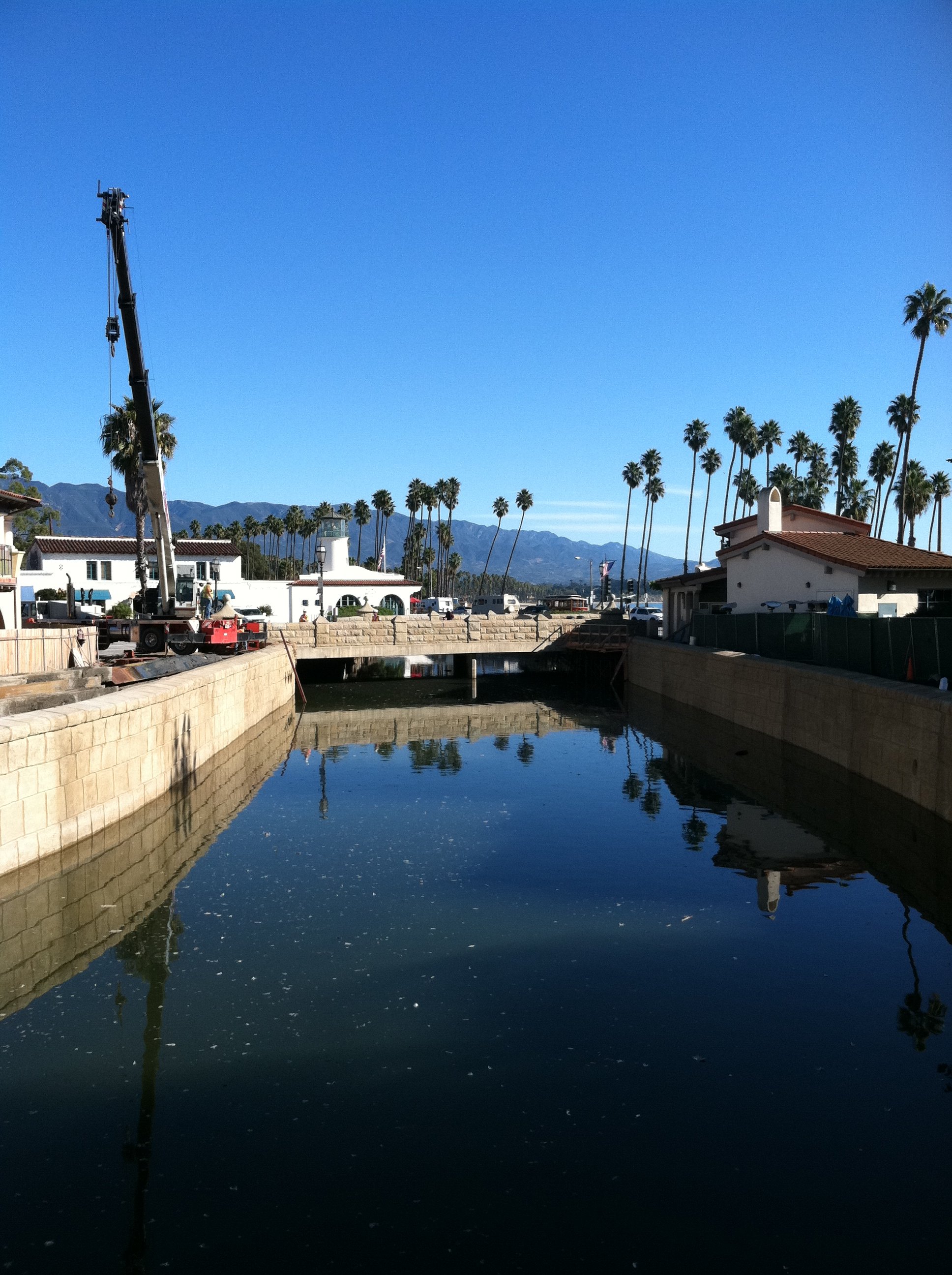 Construction crane near a canal with buildings and palm trees, mountains in the background on a clear day.