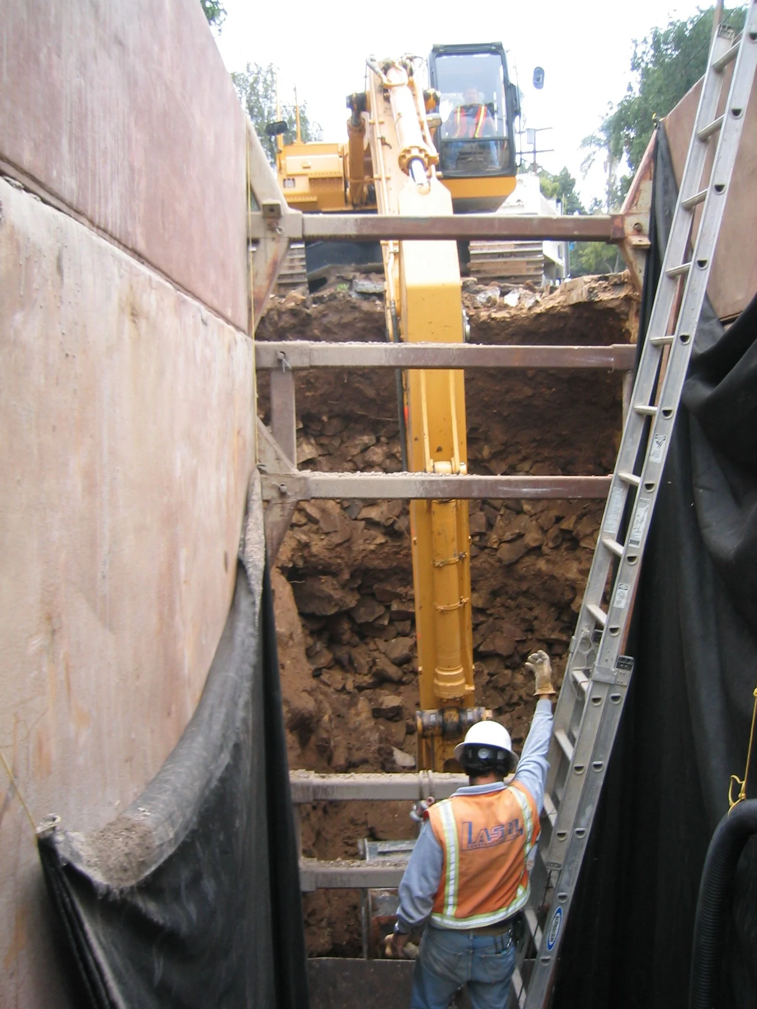 Construction workers and heavy machinery operating in a large excavation site, with a worker in protective gear and a ladder visible.