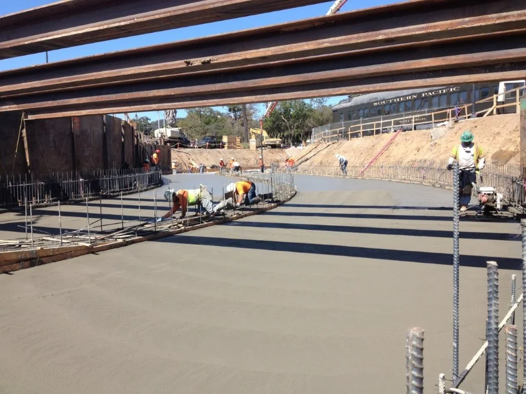 Construction workers pouring and smoothing concrete for a new road beneath a bridge, with steel rebar and construction equipment visible.