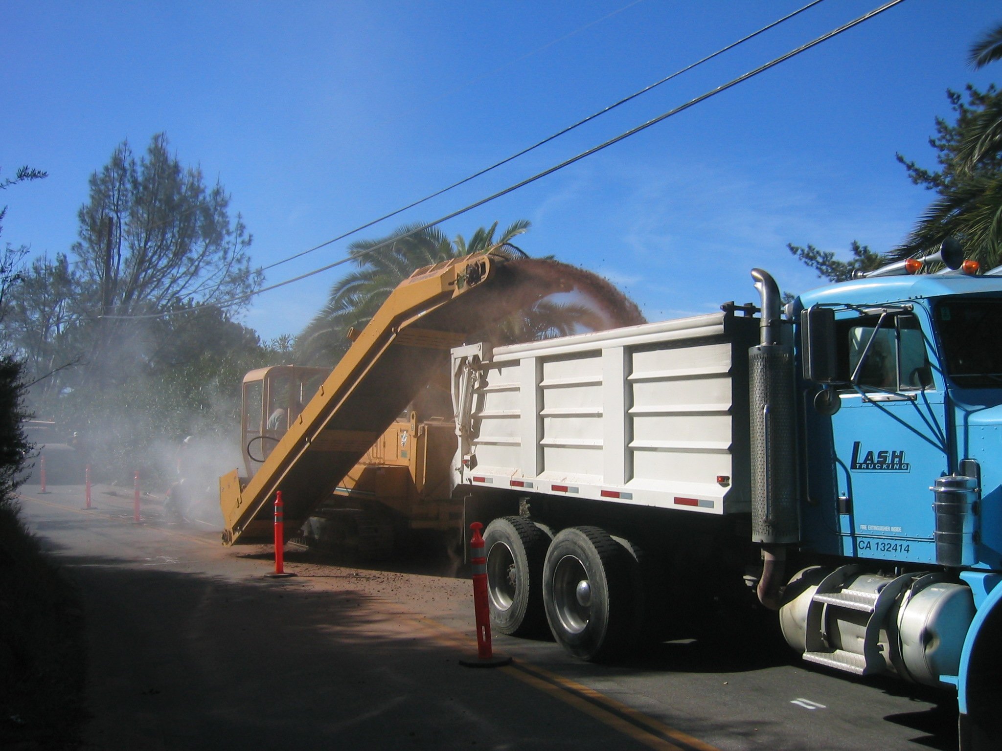 A large blue truck with a white cargo bed is on a street, with orange traffic cones and orange construction poles around it. The truck has a hydraulic lift with a large yellow garbage compactor attachment, which is unloading debris into the truck. Th