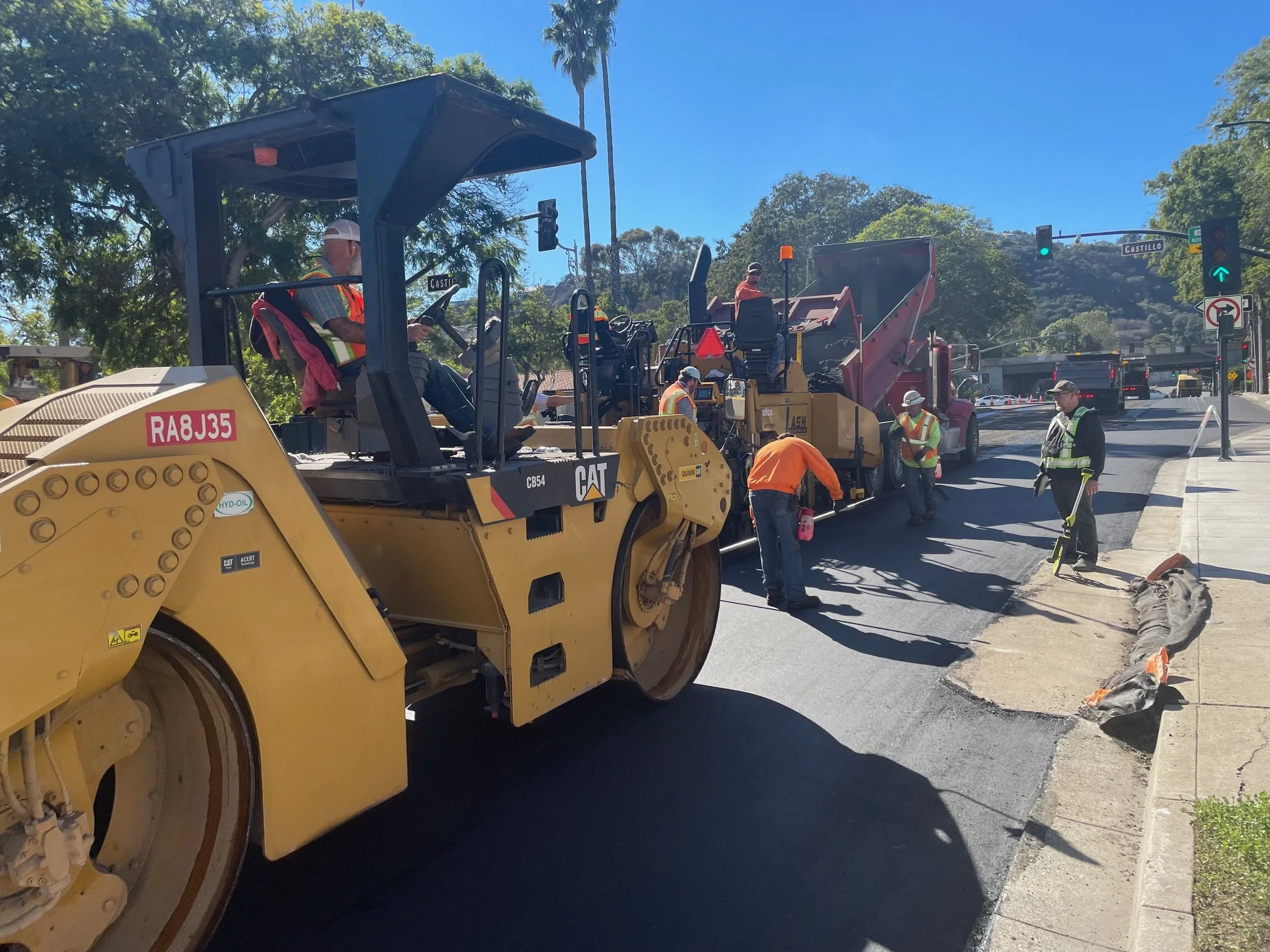 Construction workers operating heavy machinery on a city street with traffic signals and trees in the background.