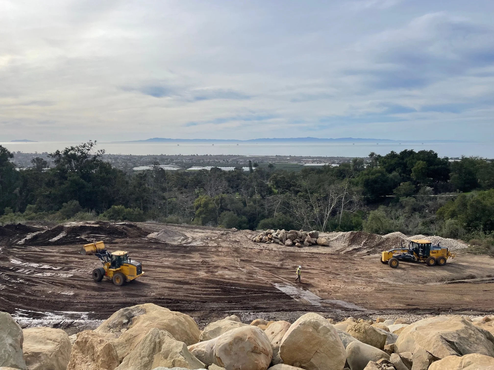 Construction site with large rocks in the foreground, bulldozers, and workers on muddy ground. In the background, there are trees, a distant city, and water with boats under a cloudy sky.