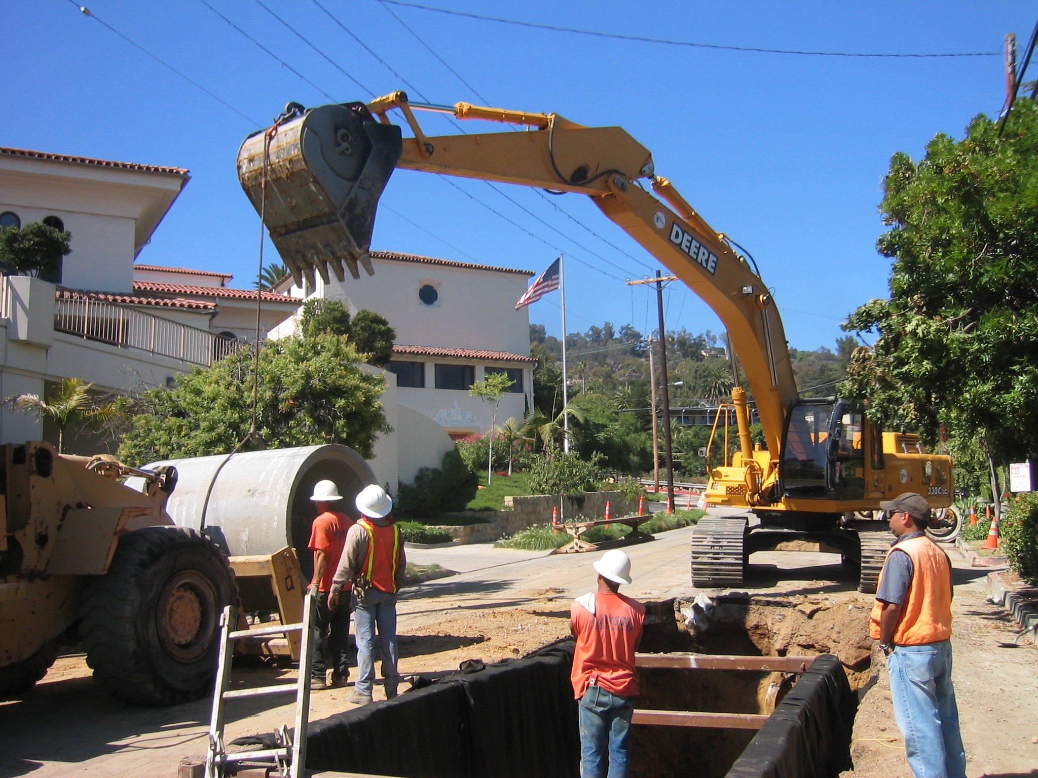 Construction workers in orange vests and white helmets working on a street with heavy machinery, including a large yellow excavator and a large truck, under a blue sky with residential buildings nearby.
